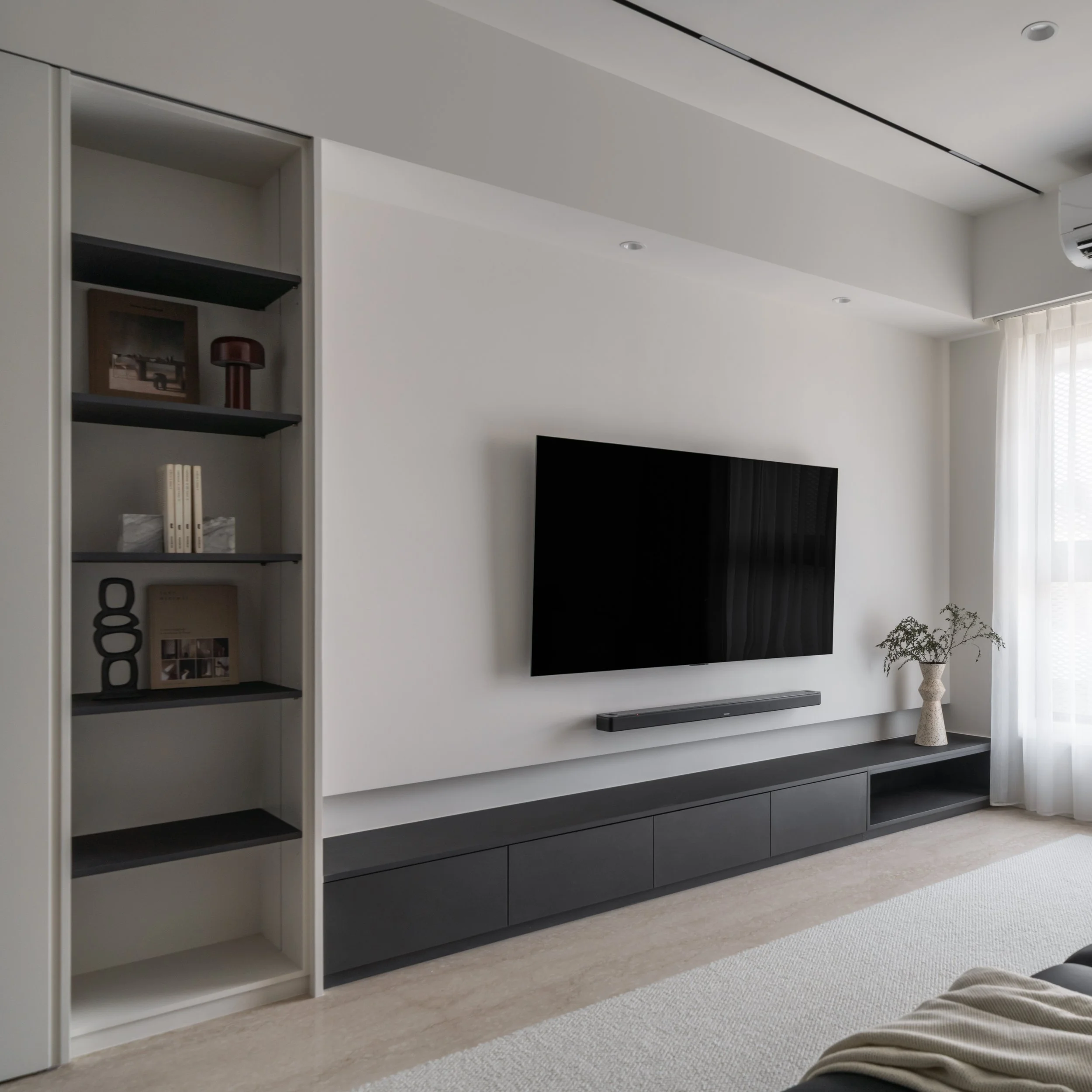 Modern living room with a large flat-screen TV mounted on white wall, black soundbar below, and open shelving unit to left with books and decorative items. Windows with sheer curtains let in natural light.