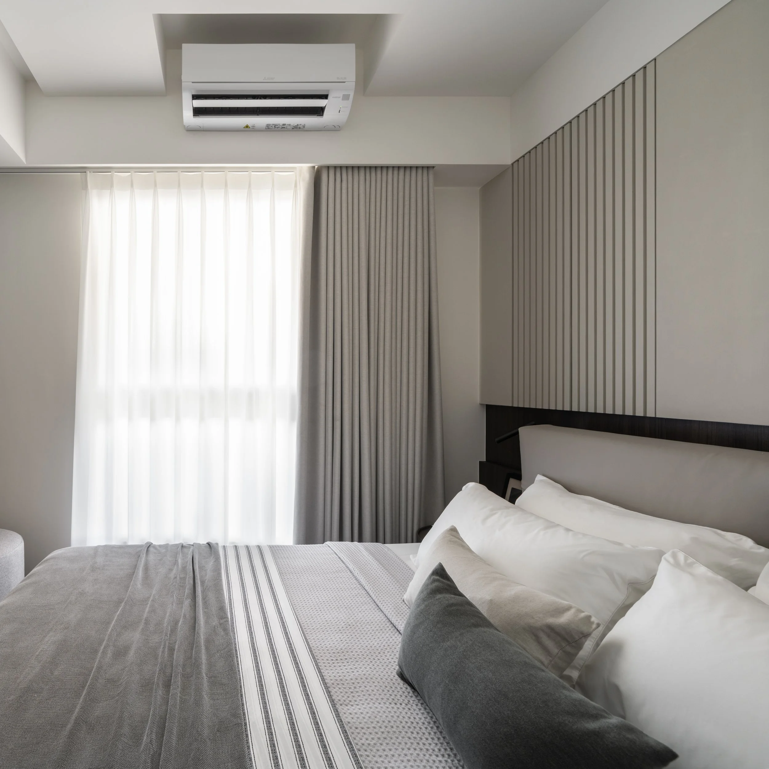 A bedroom with a bed, bedside table, light-colored curtains, and an air conditioning unit on the ceiling