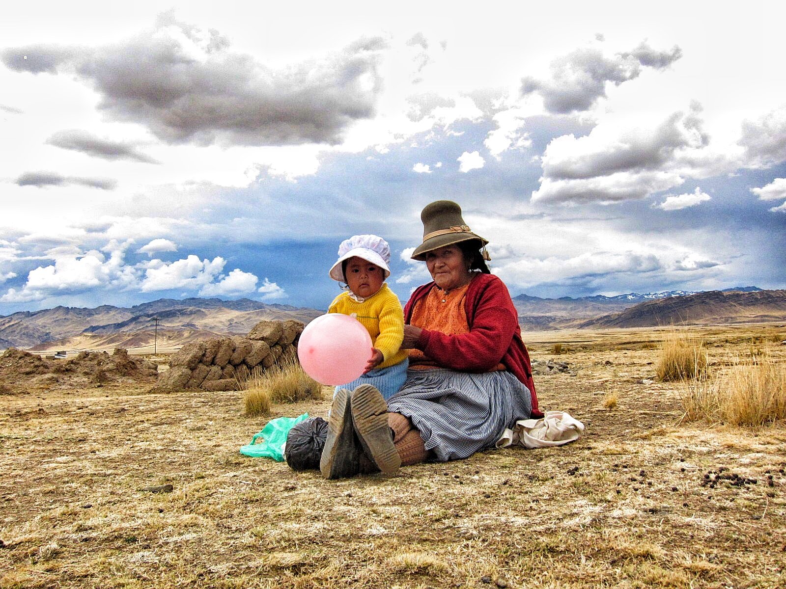 An elderly woman and a young girl sitting on the ground in a vast open landscape with mountains and cloudy sky in the background. The woman is wearing a hat, red sweater, and striped skirt, while the girl is holding a pink balloon and wearing a white