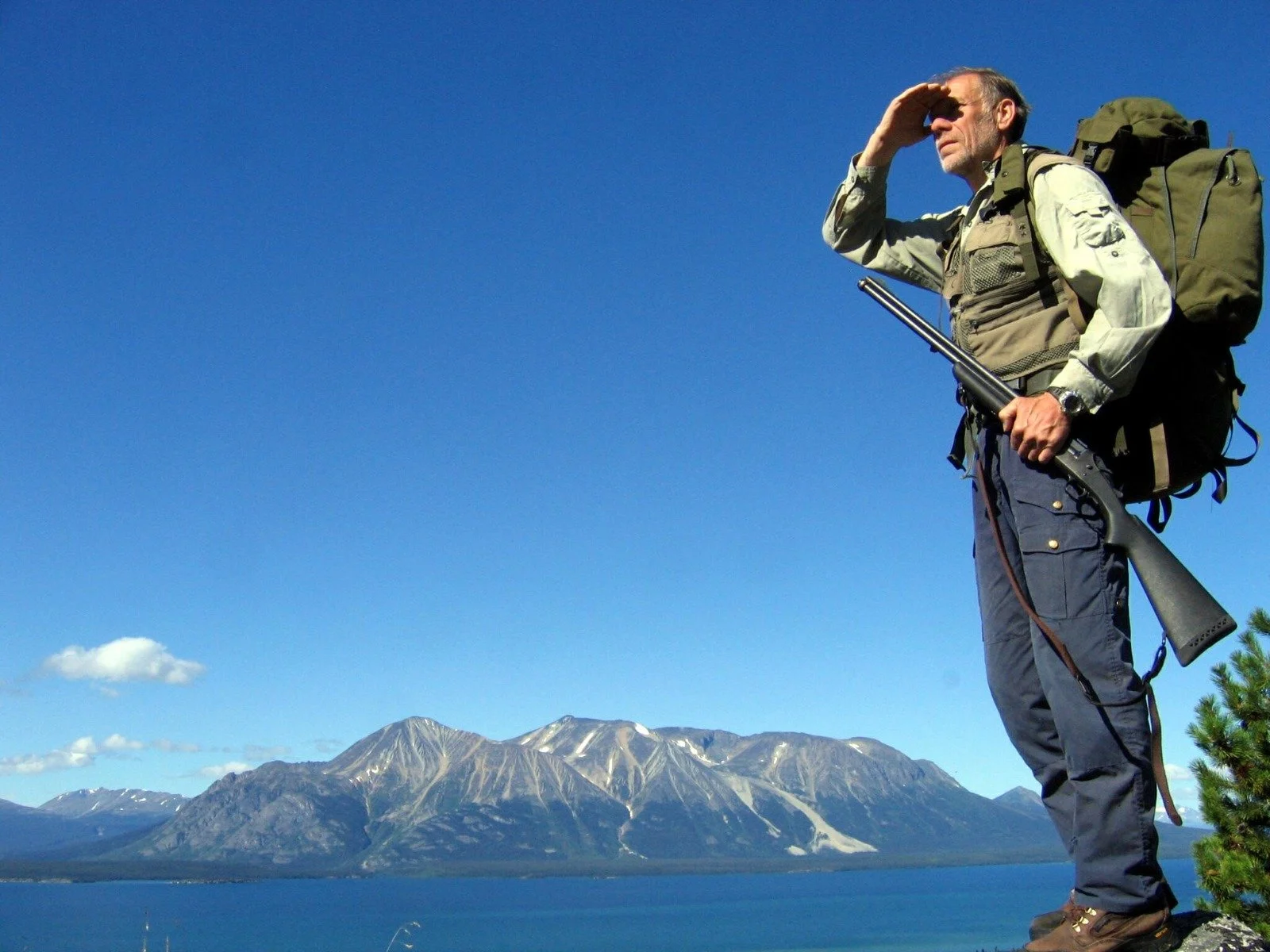 A man standing outdoors on a mountain with a backpack, holding a rifle, and looking into the distance under a clear blue sky with mountains and water in the background.
