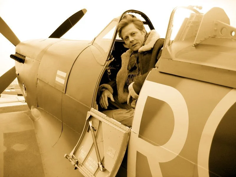 A man sitting in the cockpit of a vintage military jet aircraft, looking at the camera, with the aircraft's propeller visible in the foreground.