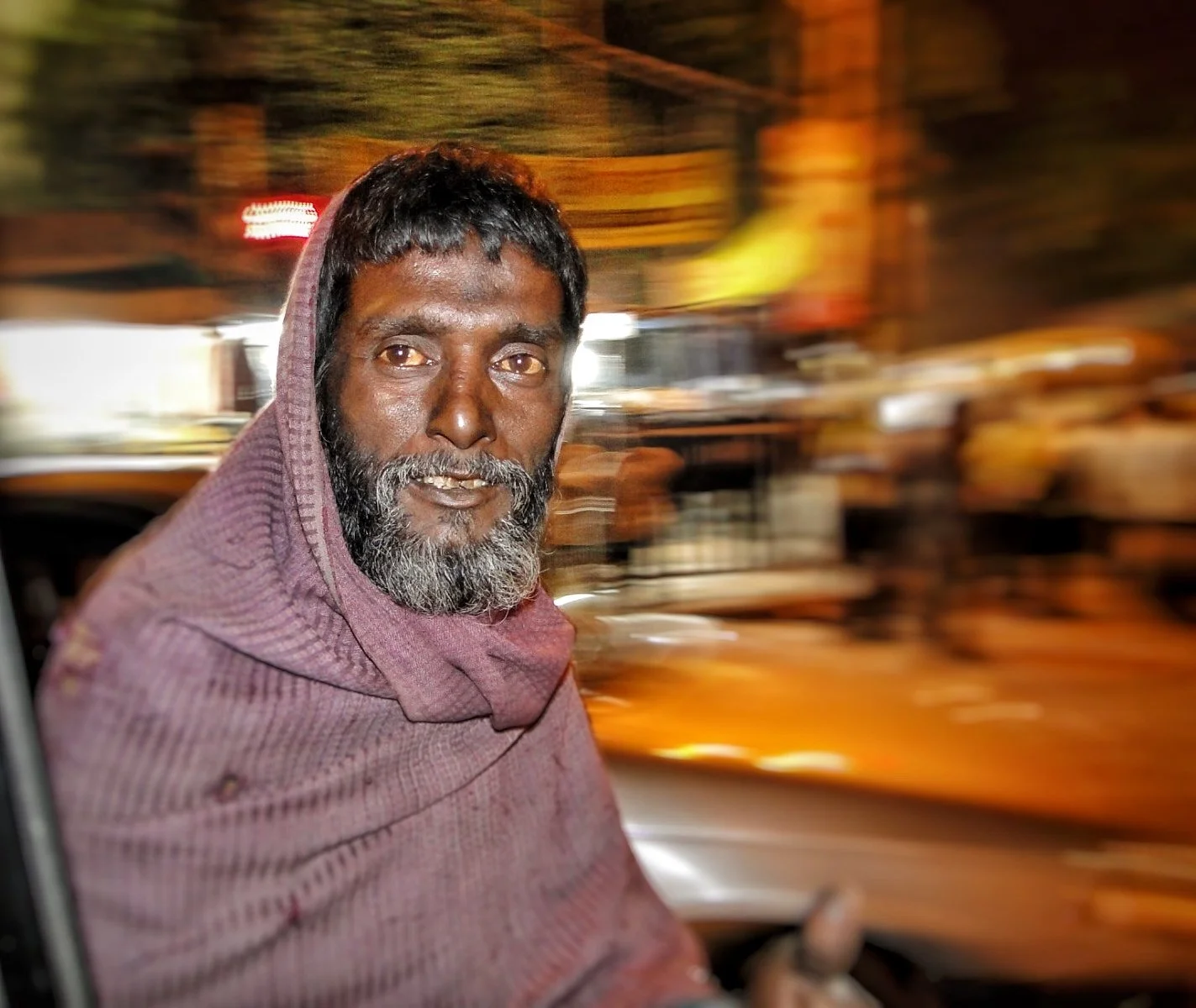 A man with a beard and mustache wearing a hoodie sitting in a vehicle at night, with a blurred city street background.