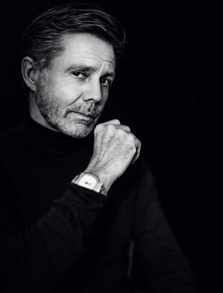 Black and white portrait of a middle-aged man with gray hair and beard, wearing a black turtleneck and a wristwatch, resting his chin on his hand against a dark background.