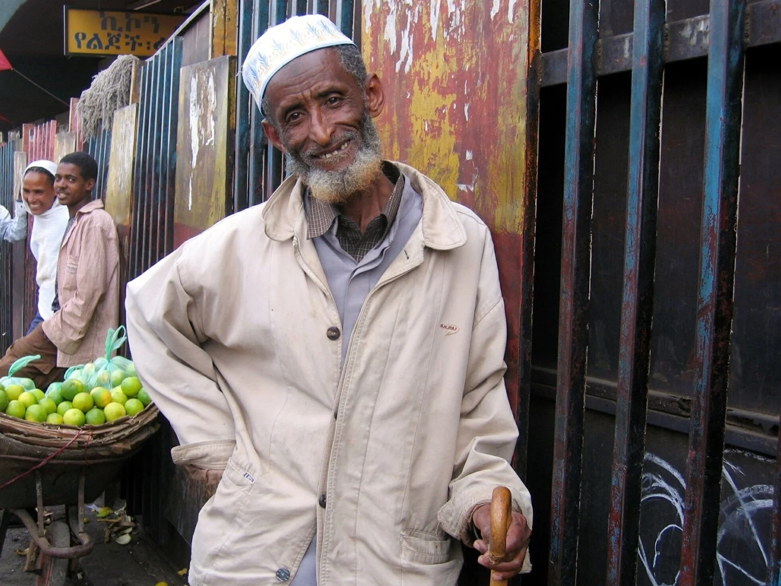 An elderly man with a beard smiling at the camera, wearing a traditional white cap and a beige jacket, standing next to a cart with green lemons in a street market.