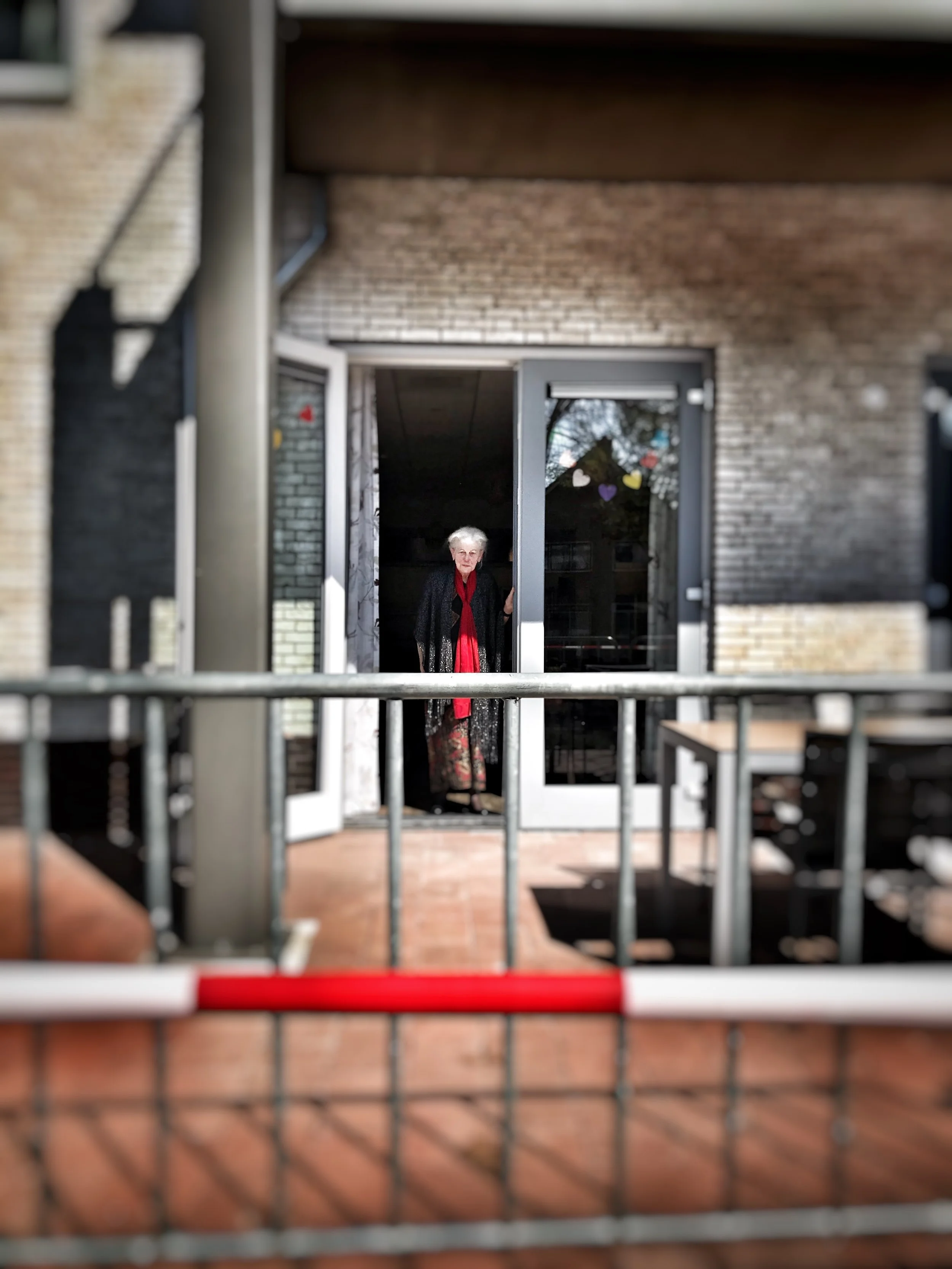 An elderly woman with white hair and glasses standing in doorway, visible through metal bars of a barrier or gate, with a brick building in the background.