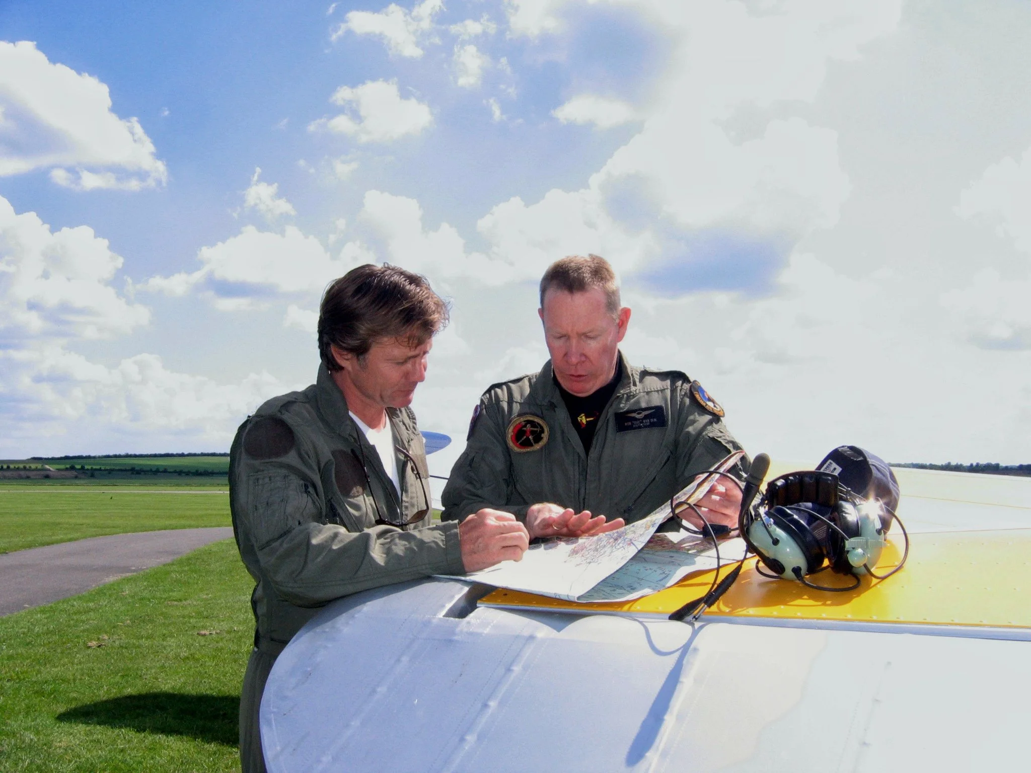 Two pilots in aviation uniforms examining a map on the wing of a small aircraft on a grassy field during daytime with partly cloudy sky.