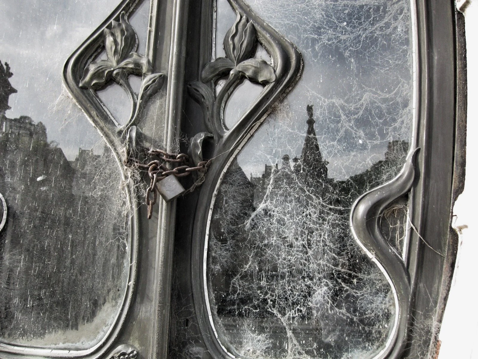 Close-up view of a locked, rusty, chained padlock on a tarnished, dusty, and cobweb-covered window or frame with reflections of buildings and a clock tower in the glass.