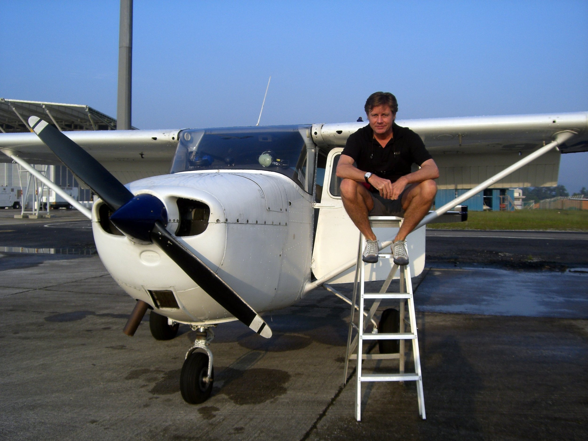 A man sitting on the wing of a small private airplane parked on an airfield. There are airport buildings and equipment in the background under a clear blue sky.
