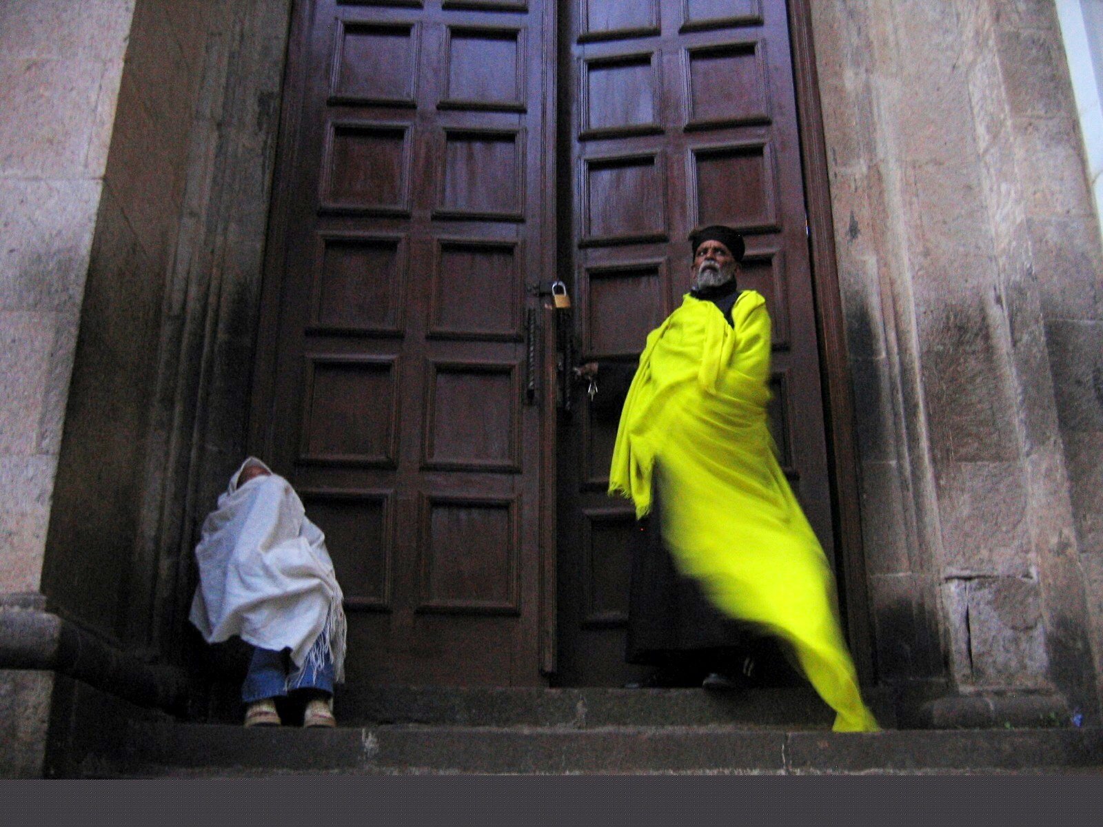 A man dressed in a bright yellow traditional garment standing in front of large wooden doors with a padlock. A person dressed in white and wearing a face mask is sitting on the steps next to him, partially visible.