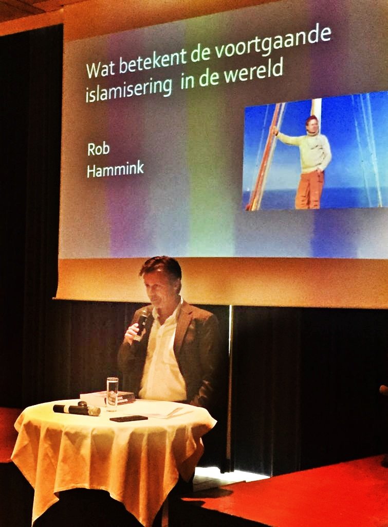 A man in a suit holding a microphone stands at a round table with a glass of water, papers, and a remote control, in front of a large screen displaying a presentation titled 'What does the ongoing Islamification in the world mean,' by Rob Hammink.