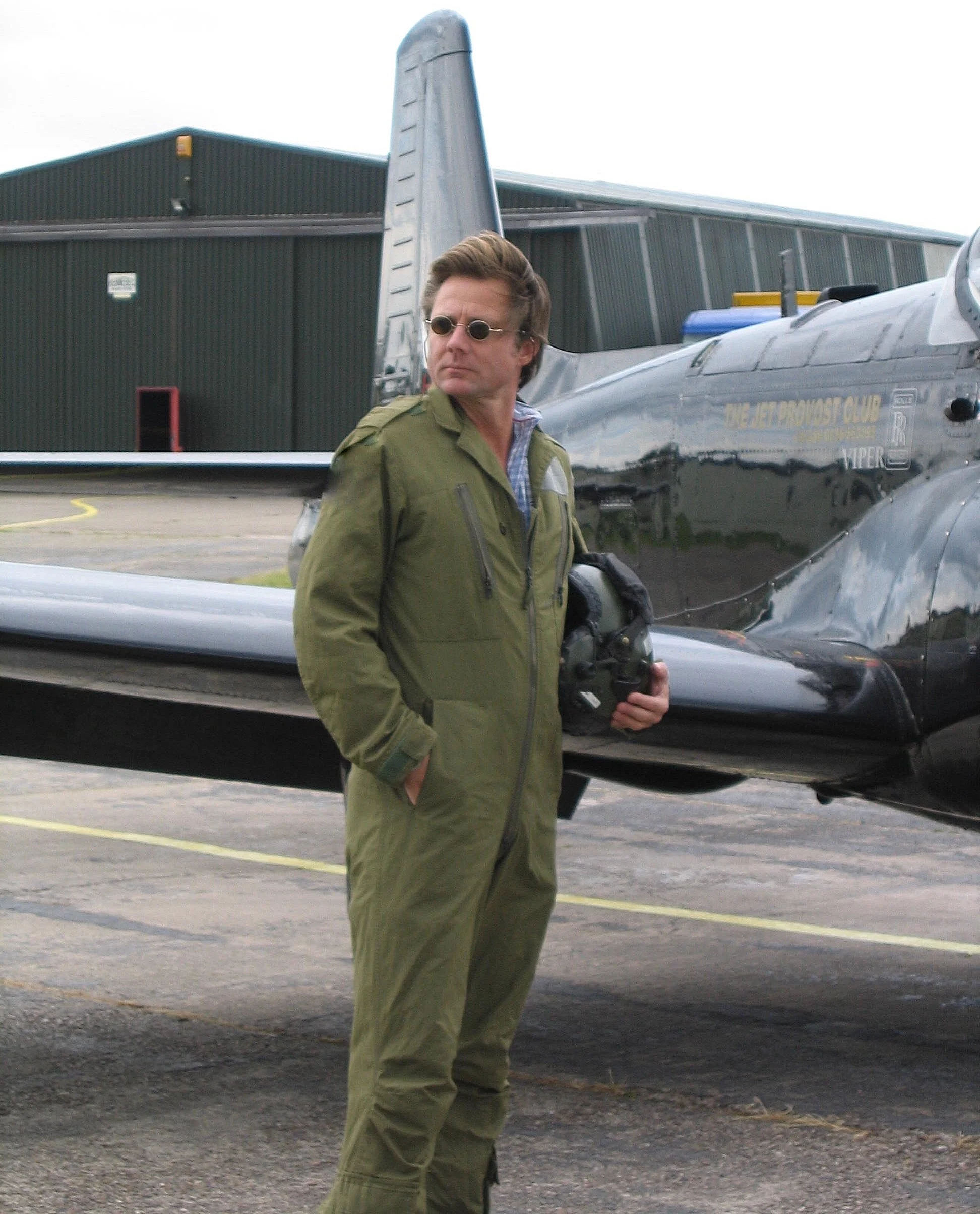 Man wearing military-style green jacket with sunglasses standing near a fighter jet at an airfield.