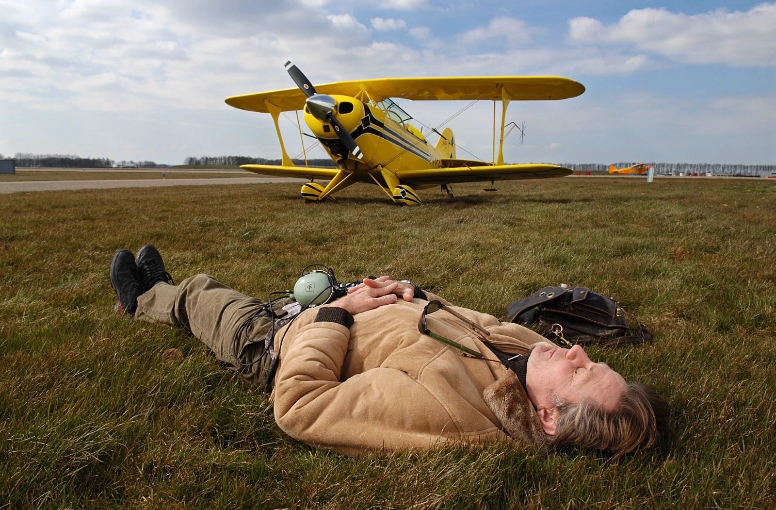 A man lying on the grass with a yellow biplane in the background on an overcast day.