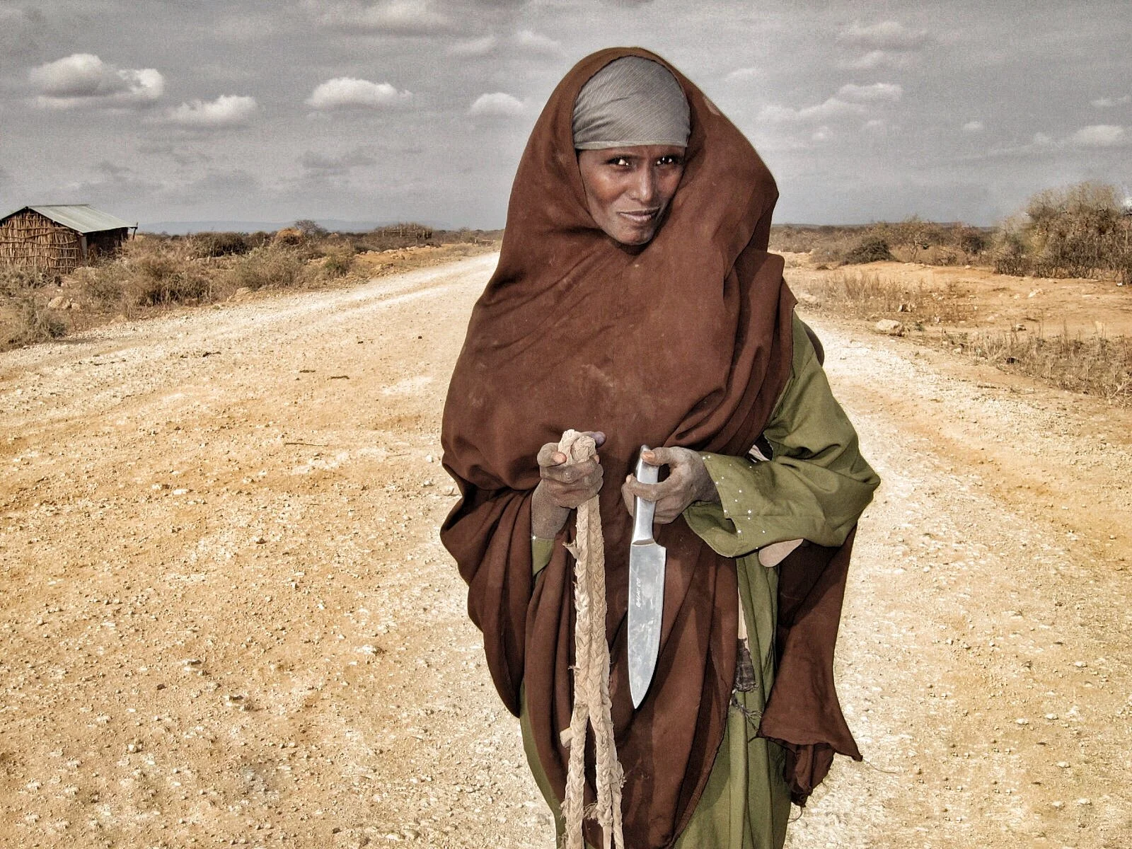 A woman dressed in traditional clothing holding a knife and a rope standing on a dirt road in a rural, arid landscape with sparse vegetation and a small wooden structure in the background.