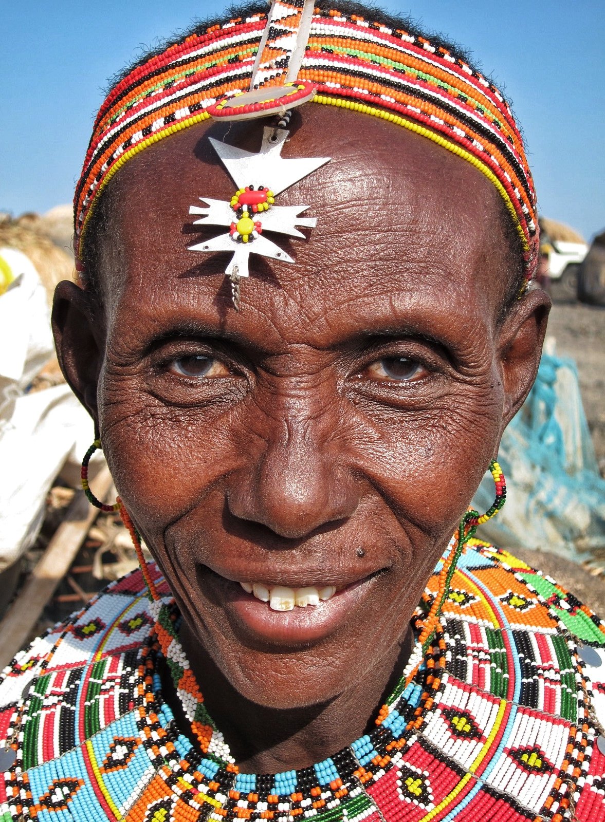 Close-up of an elderly woman wearing traditional colorful beaded jewelry and a beaded headband, smiling outdoors with a clear blue sky in the background.
