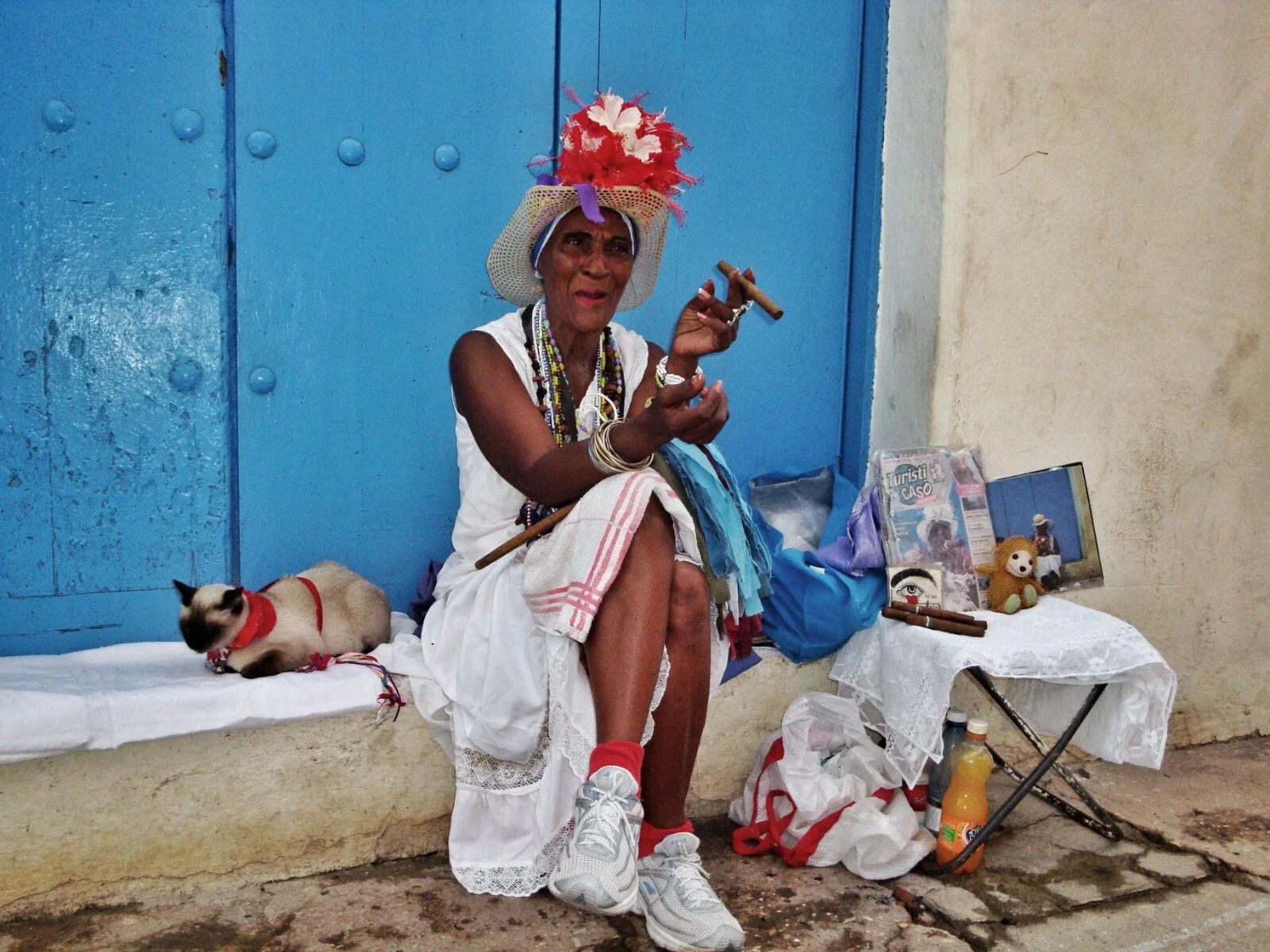 An elderly person with dark skin sitting on a white cloth, wearing a large, colorful feathered hat, white dress, and white sneakers, holding a cigar. To their left, a Siamese cat resting on a small bed, and to their right, a small makeshift table wit