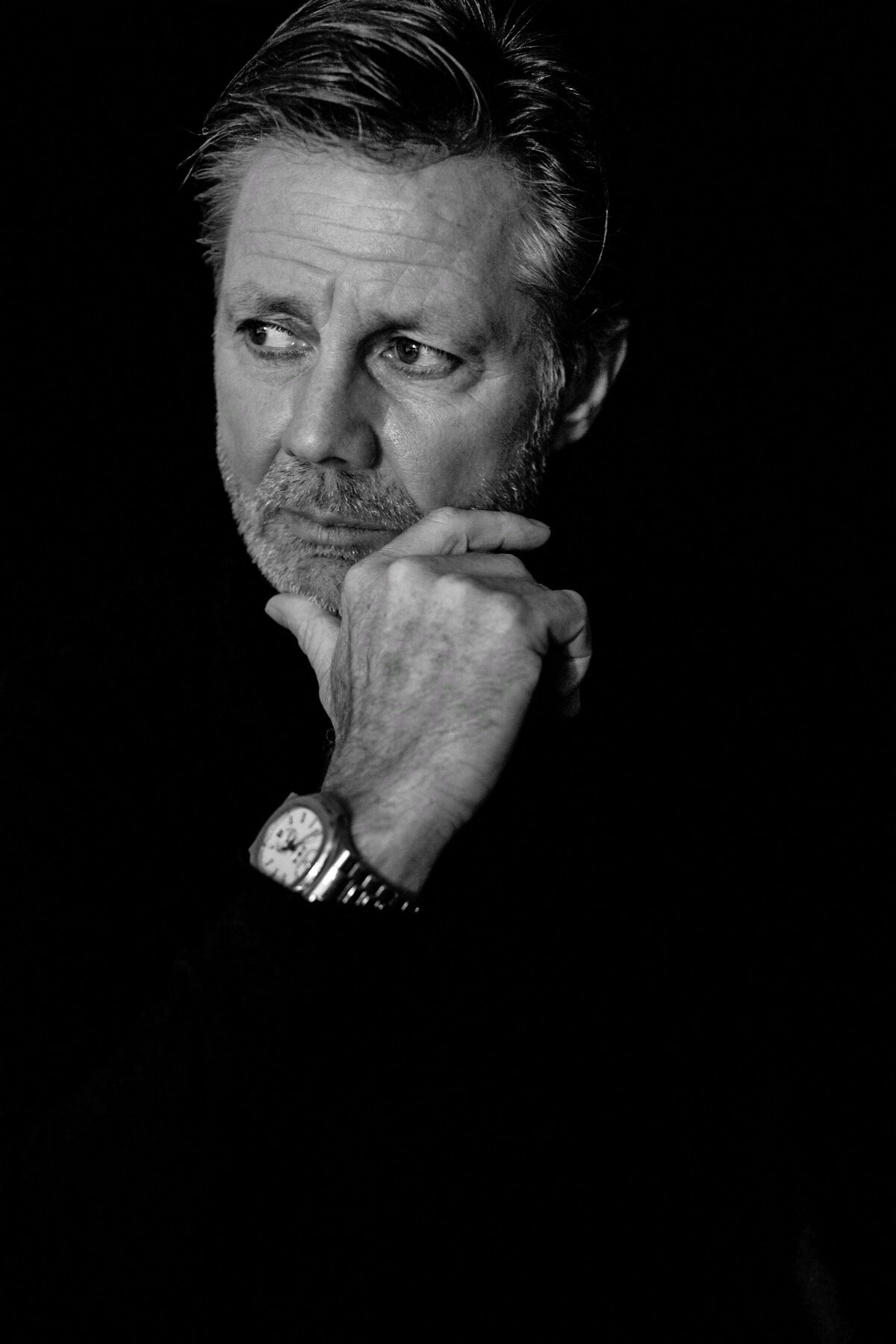 Black and white portrait of an older man with gray hair and a beard, looking thoughtfully to the side with his hand on his chin, wearing a watch on his left wrist against a dark background.