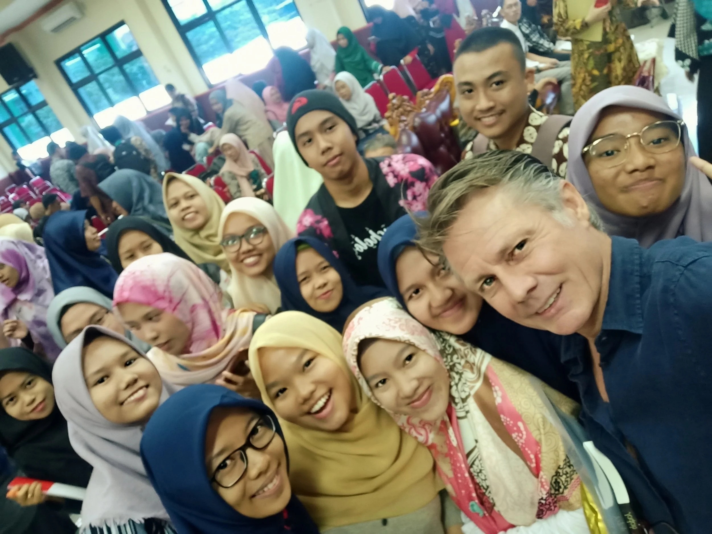 Group of smiling children and adults taking a selfie in a crowded indoor space.