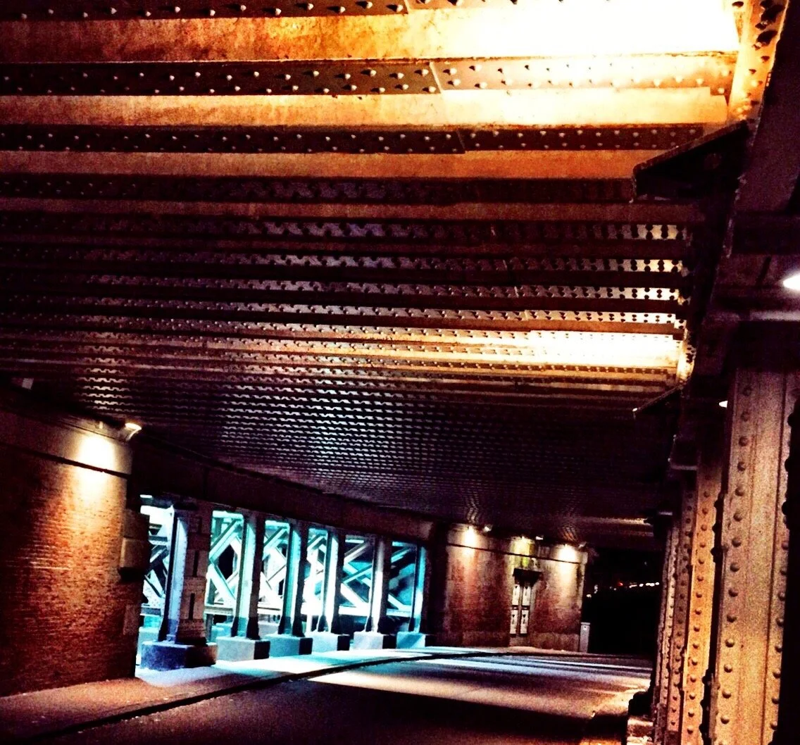 Under a bridge with rusted metal beams and rivets, a brick wall and illuminated structural supports are visible. Light filters through the gaps, casting shadows on the ground.