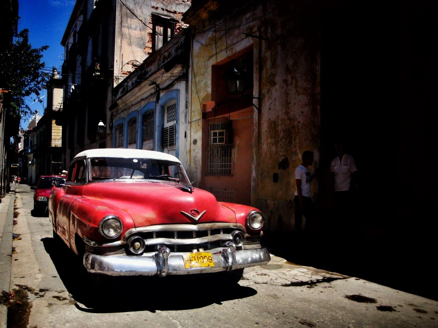 A vintage red and white car parked on a narrow street with old buildings and two people standing on the sidewalk, in a sunny urban scene.
