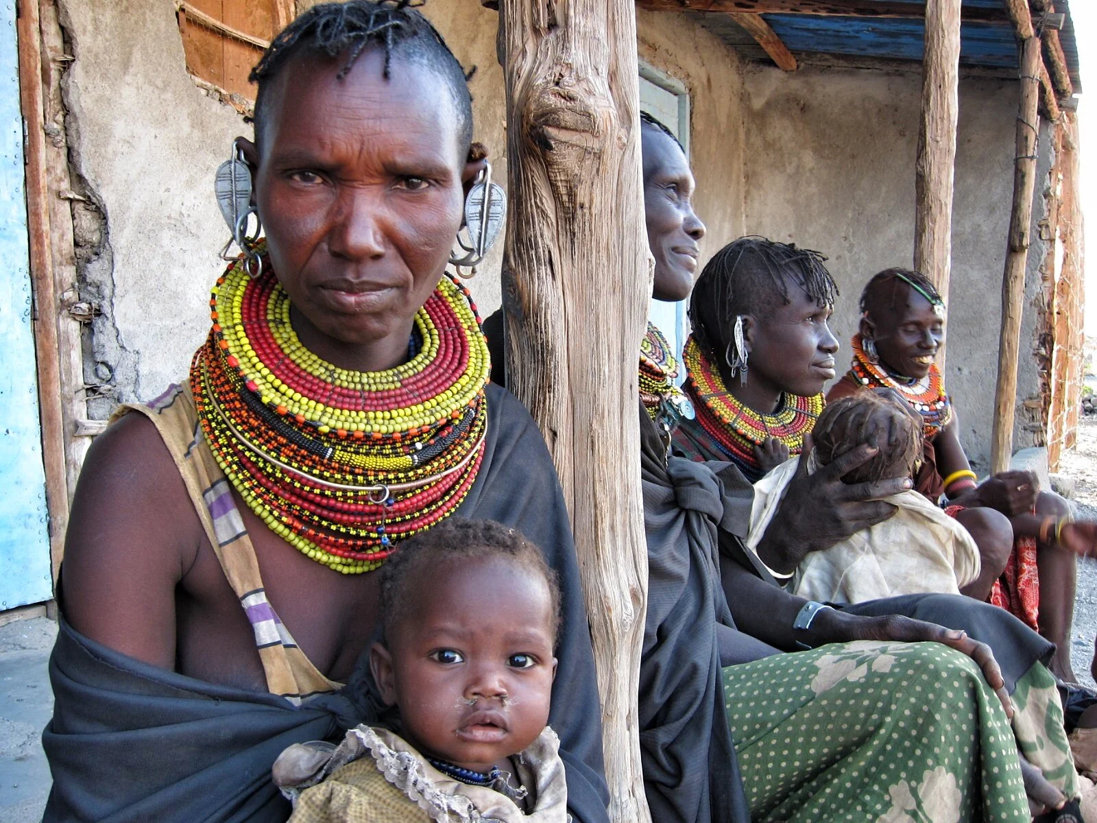 A group of women and children sitting outside, wearing traditional jewelry and clothing, with some women wearing multiple layers of beaded necklaces.