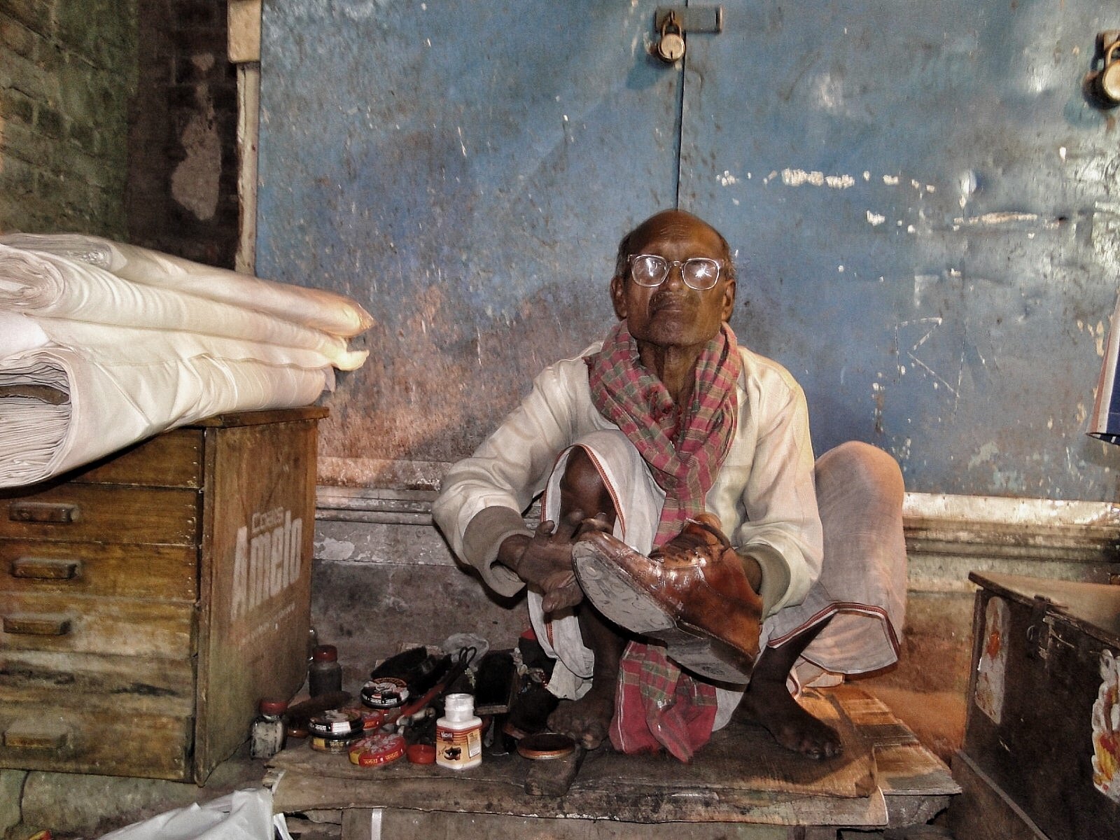 An elderly man wearing glasses and a worn scarf sitting on the floor in a modest room. He is holding a shoe and appears to be polishing or repairing it. The room has a blue painted wall with peeling paint, a wooden chest, some shoes, and jars scatter