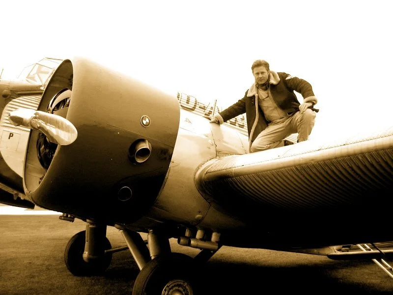 A person crouching next to a vintage aircraft, posing on a wing.
