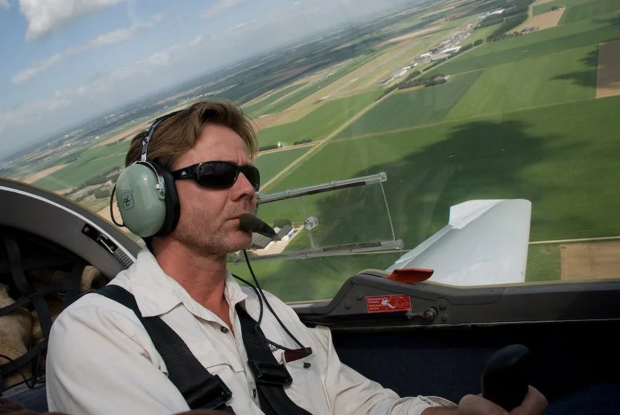 A man flying an airplane, wearing sunglasses, headphones, and a white shirt, with farmland visible through the cockpit window.