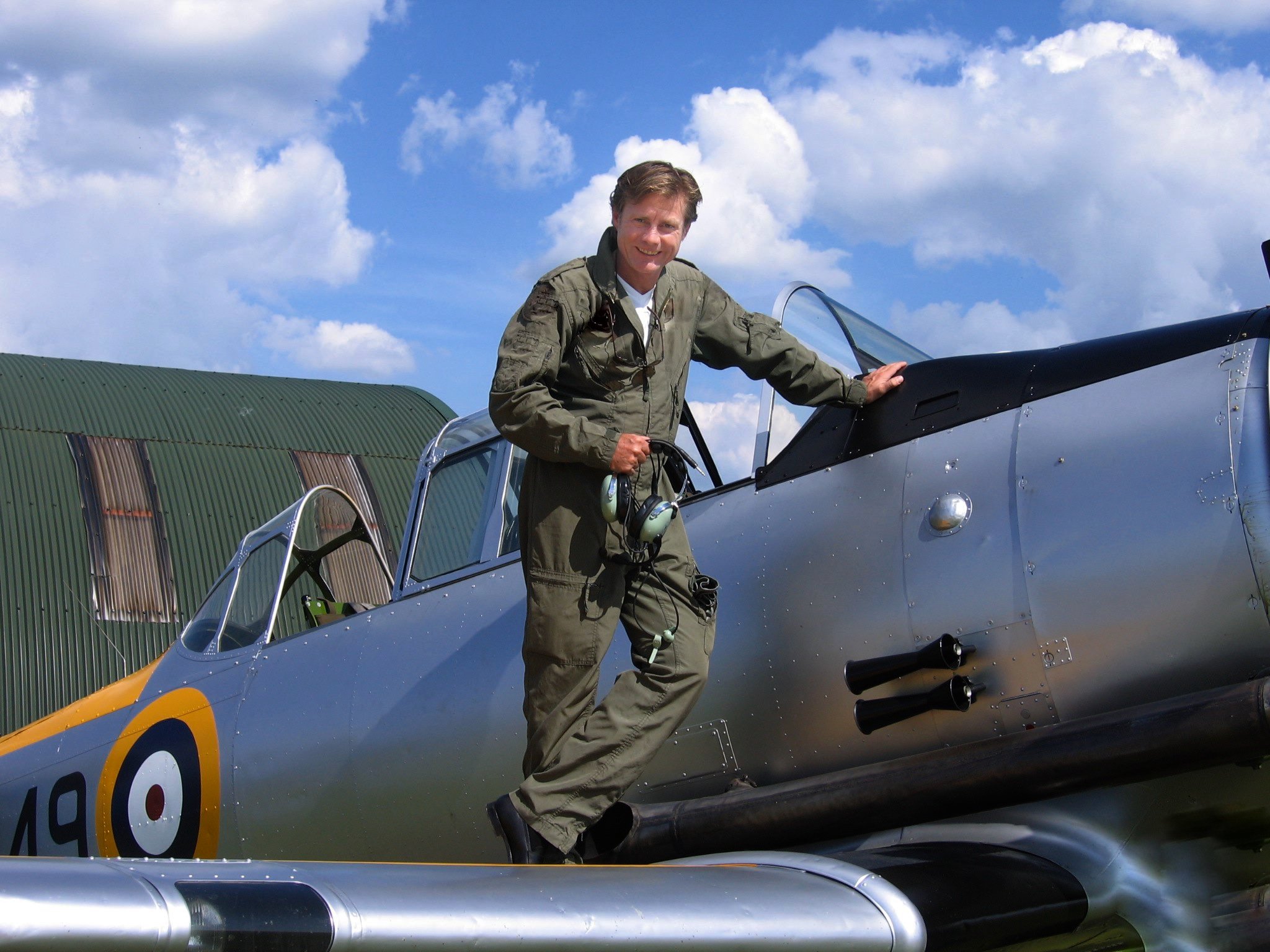 A man in a flight suit standing on an aircraft, smiling, with a cloudy sky and hangar in the background.