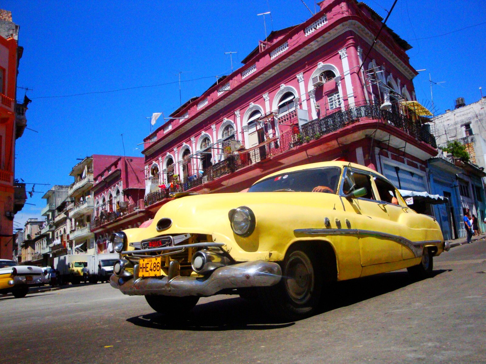 A yellow vintage car driving on a street in front of colorful buildings with pink, white, and blue facades, under a clear blue sky in a lively city scene.