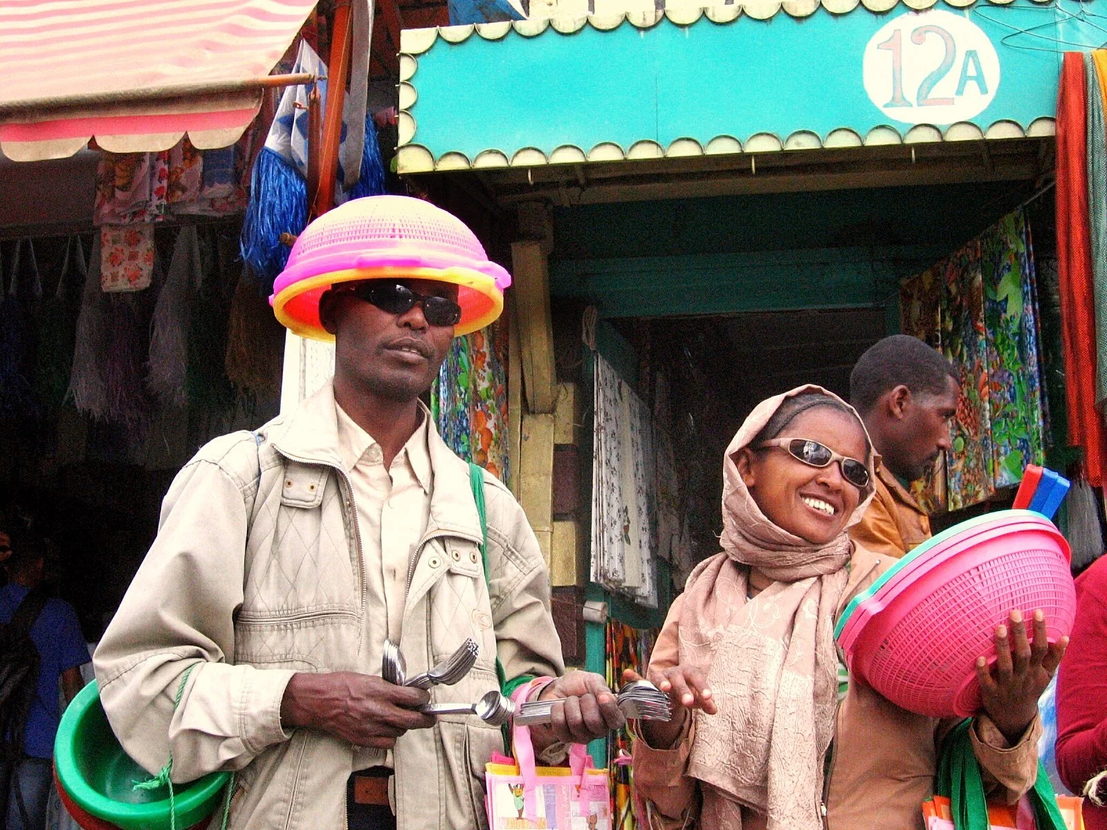 Three people standing in front of a market stall, one man wearing sunglasses and a hat made of colorful plastic baskets, a woman smiling with sunglasses and headscarf, holding pink plastic baskets, and another man looking away holding a blue item, co