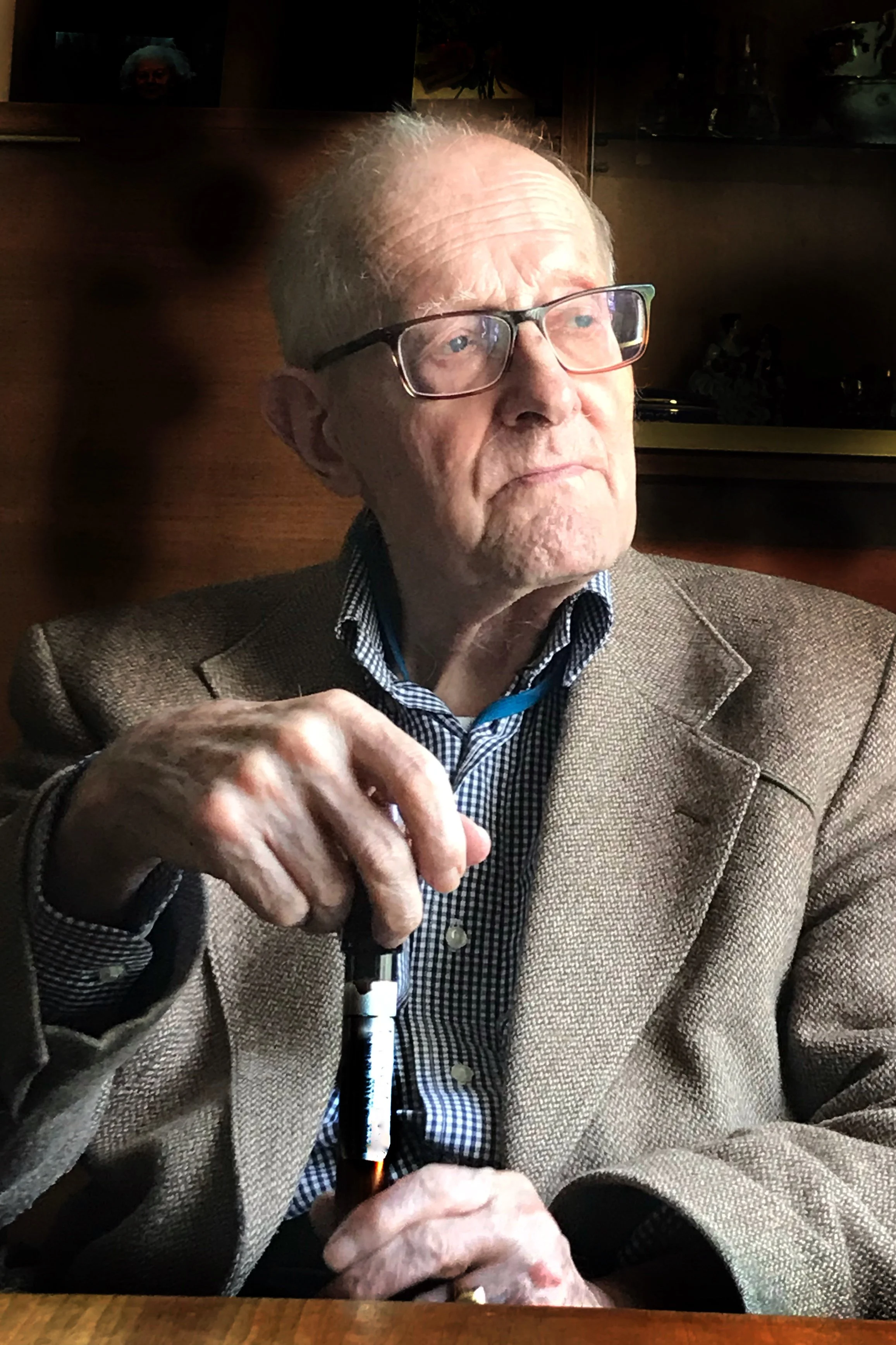 An elderly man with glasses and white hair, wearing a brown blazer and a checkered shirt, sitting at a table with a dark background. He appears to be in a restaurant or cafe, holding a microphone in his right hand.