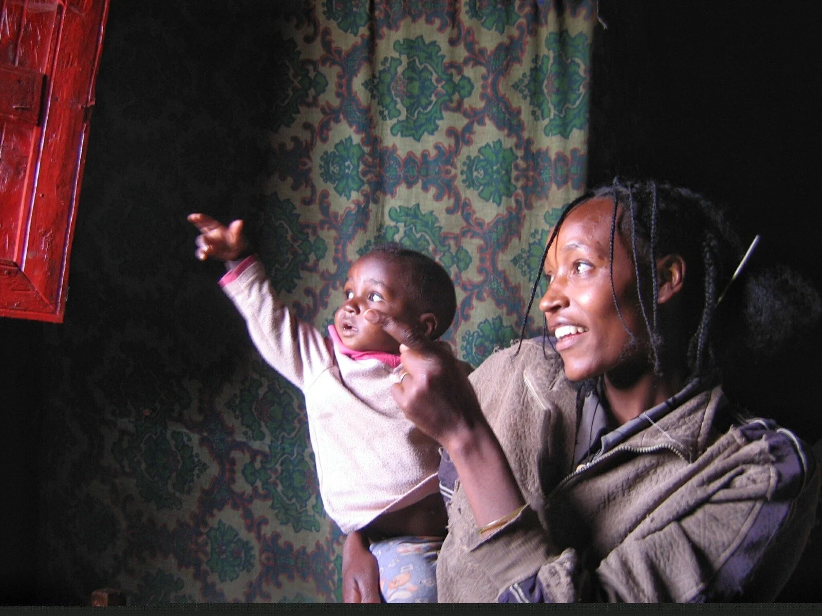 A woman and a young child sitting indoors near a colorful, patterned wall hanging. The woman is smiling and pointing at something outside the frame, while the child is reaching out with one hand and touching her face with the other. The scene is warm