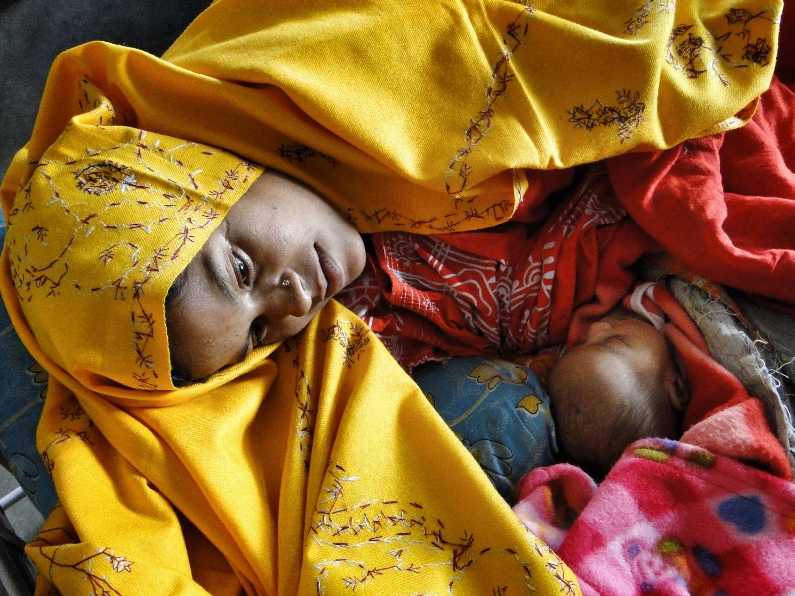 A woman lying down with a small child or baby close to her, both covered with colorful blankets. The woman is wearing a yellow headscarf with floral embroidery and has a nose ring. The baby is wrapped in a pink and multicolored blanket, and the scene