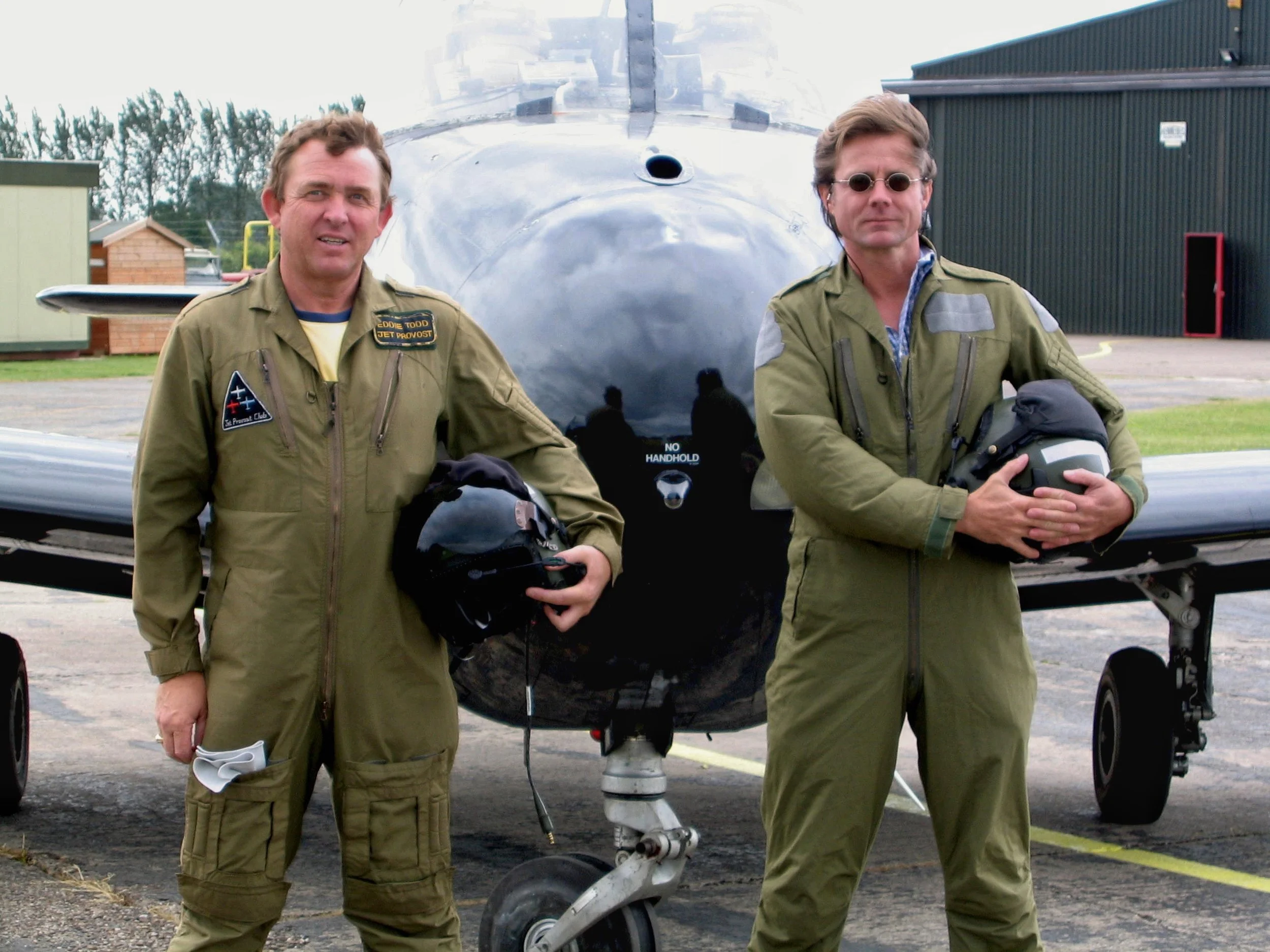 Two men in flight suits standing in front of a fighter jet on an airstrip. The man on the left is holding a helmet and some papers. The man on the right is holding a helmet with both hands, and they are posing for the photo.