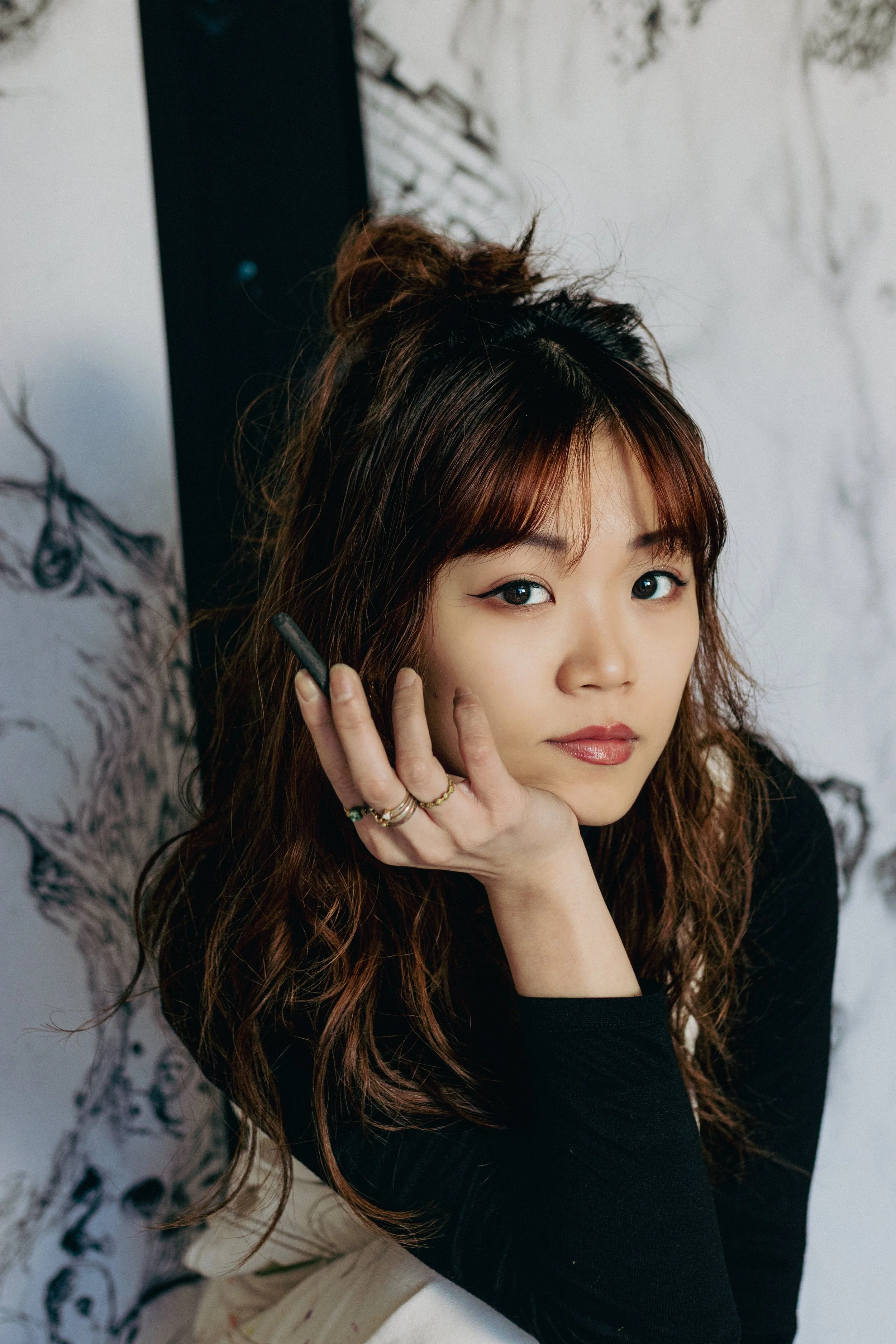 Portrait of a woman with long, wavy brown hair and bangs, wearing black eyeliner and a black top, sitting beside a wall with abstract black and white drawings.