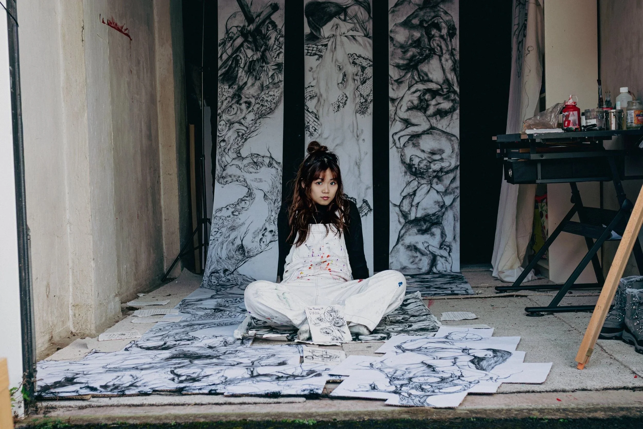An artist sits cross-legged on the floor in front of a large black and white drawing backdrop, surrounded by numerous paper sketches. The artist appears to be a young woman with dark hair styled in a messy bun, wearing a black long-sleeve shirt and white overalls splattered with paint. The background features multiple detailed sketches and drawings of faces and abstract figures.