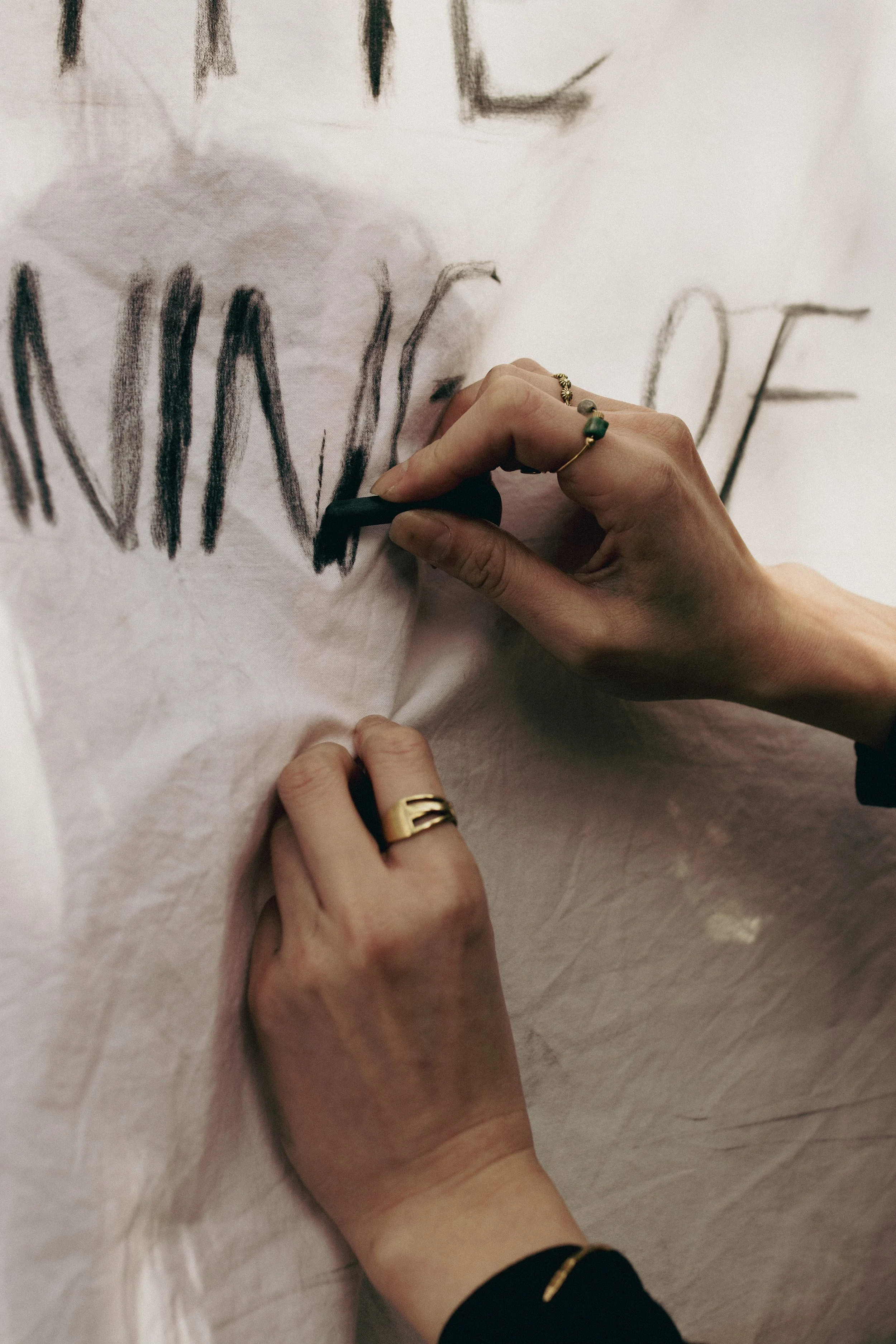 A person is using a black pastel or charcoal to draw on a white fabric or paper surface, creating large, bold, capitalized black letters.