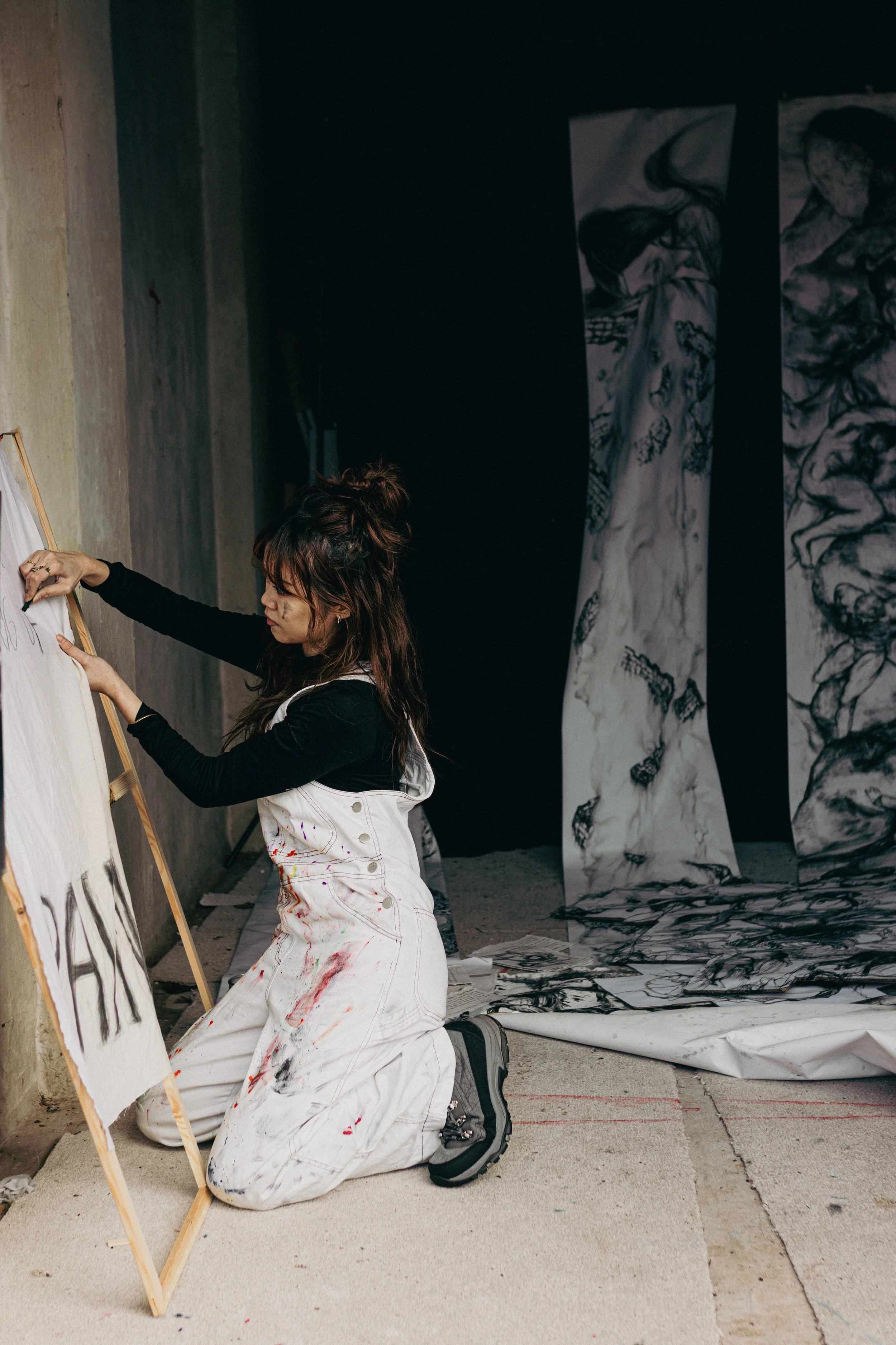 A young woman kneeling on the floor painting a sign on an easel in an art studio. She is wearing a white apron with paint stains and black shirt, and is focused on her work. Behind her are large abstract black and white paintings hanging on the wall.