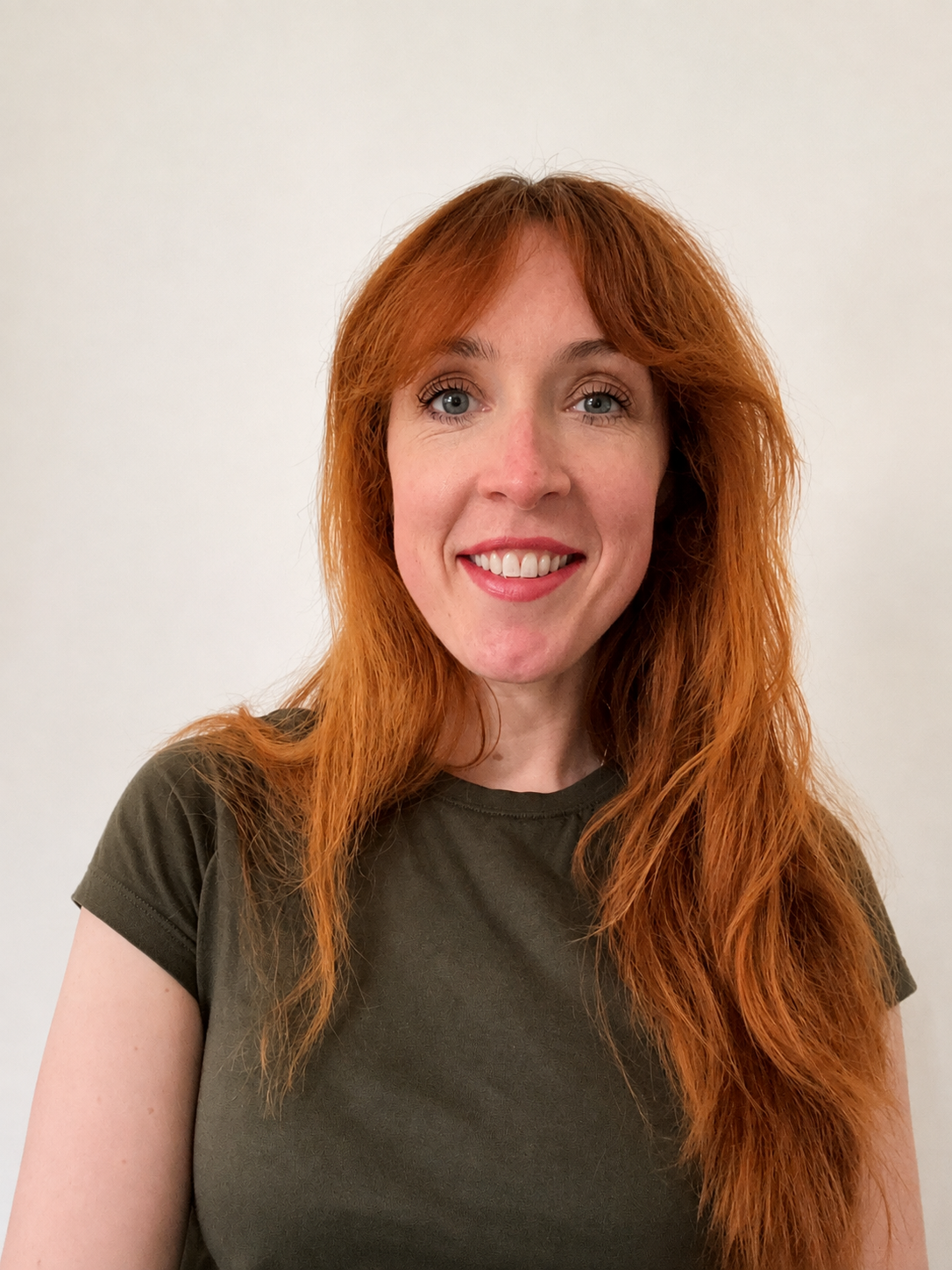 Portrait of a woman with long, wavy red hair, wearing a dark green t-shirt, smiling against a plain white background.