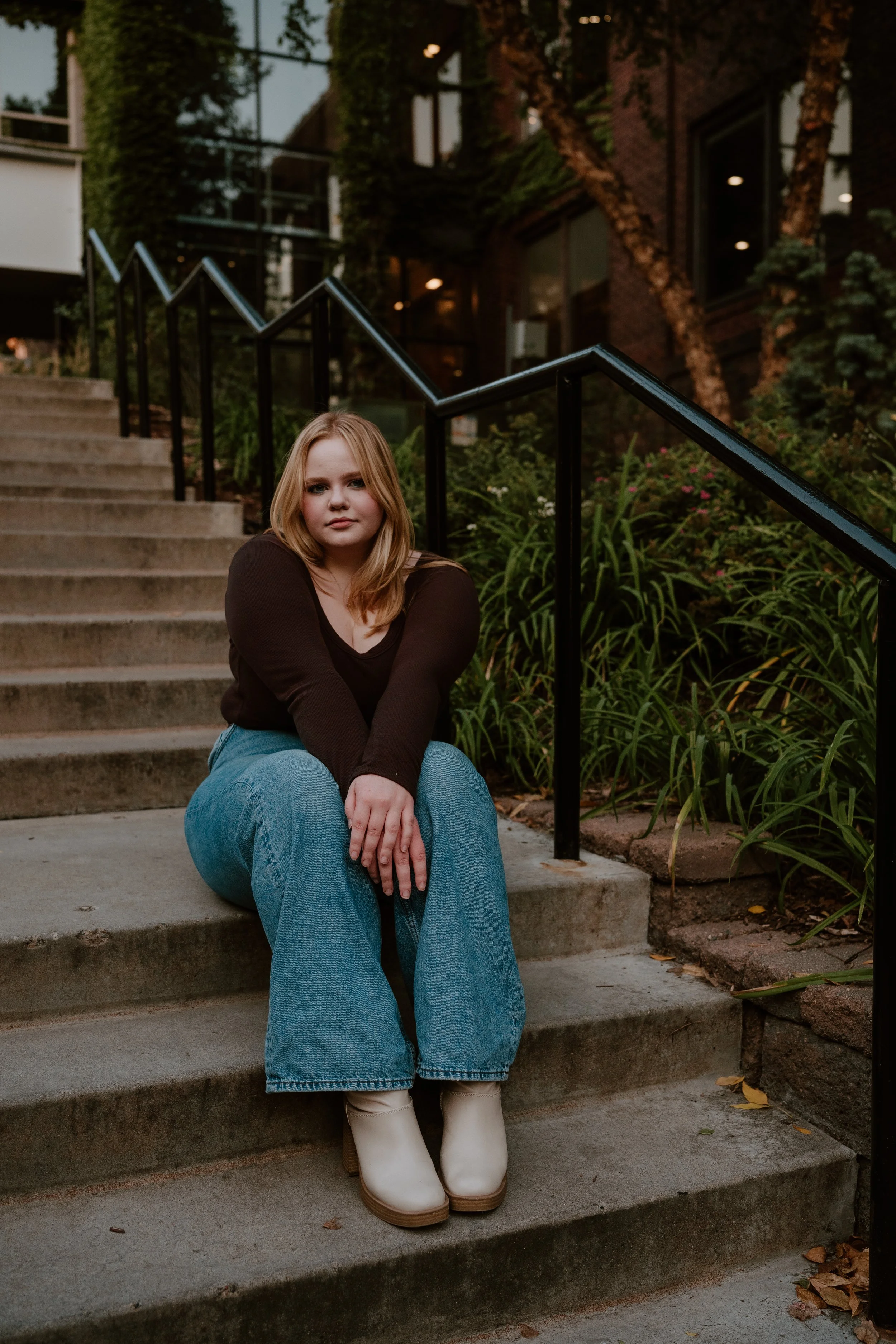 A young woman with blonde hair sitting on outdoor stairs with a railing, surrounded by greenery, at dusk in an urban setting.