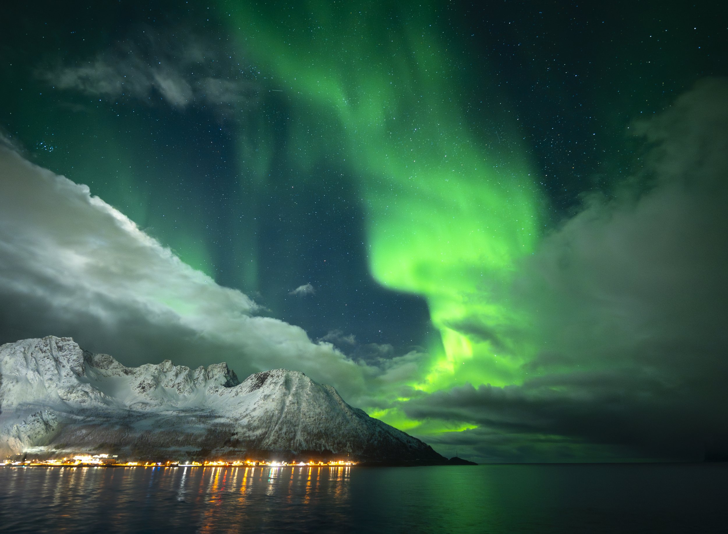 Green aurora spiralling over snow-covered peaks and a lit fishing village on the fjord, Senja, Arctic Norway