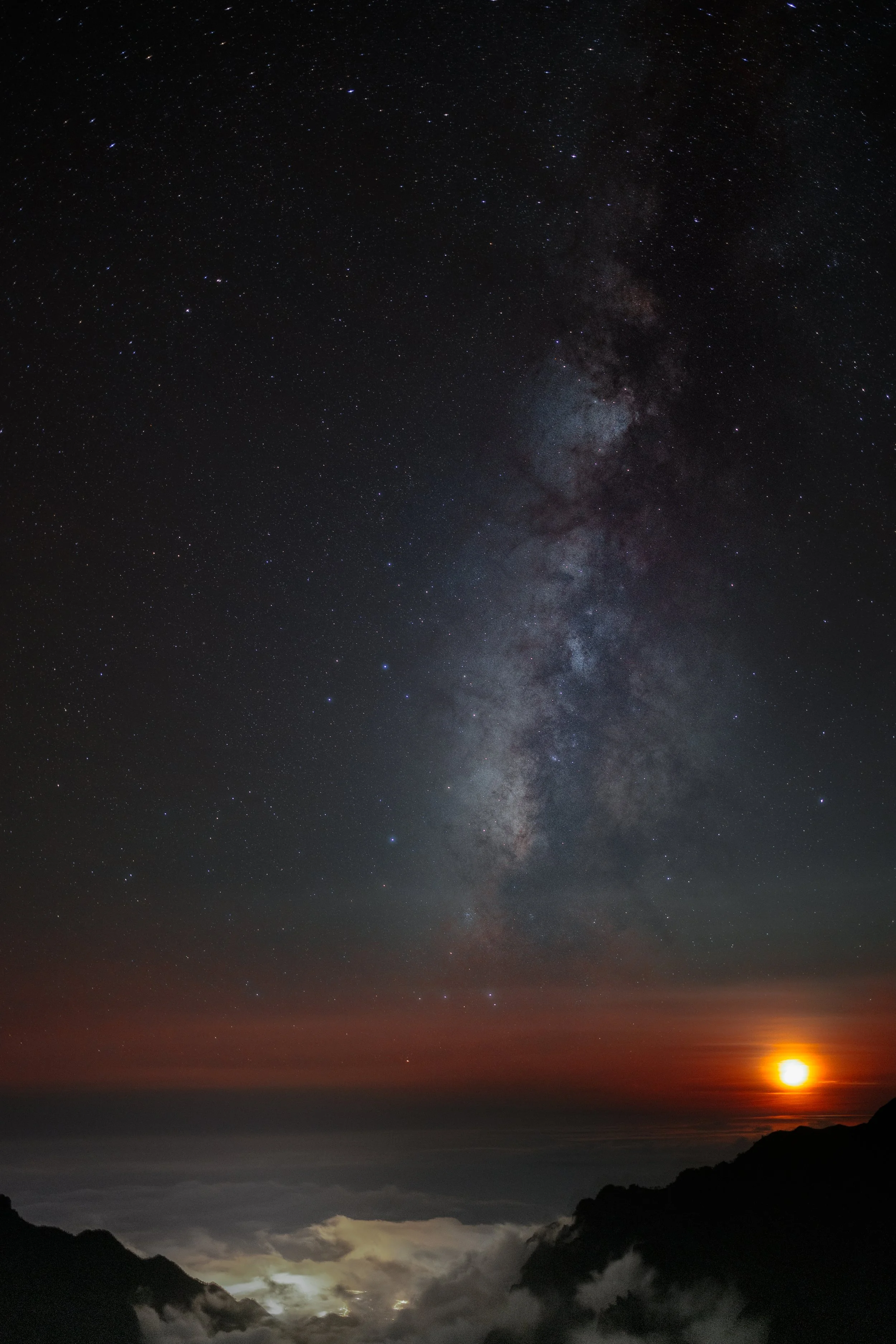 The Milky Way core above the sea of clouds from near Roque de los Muchachos, La Palma, with the moon setting orange on the horizon — the summit sits at 2,400 metres, above the trade-wind inversion that traps the cloud layer and the island's light ben