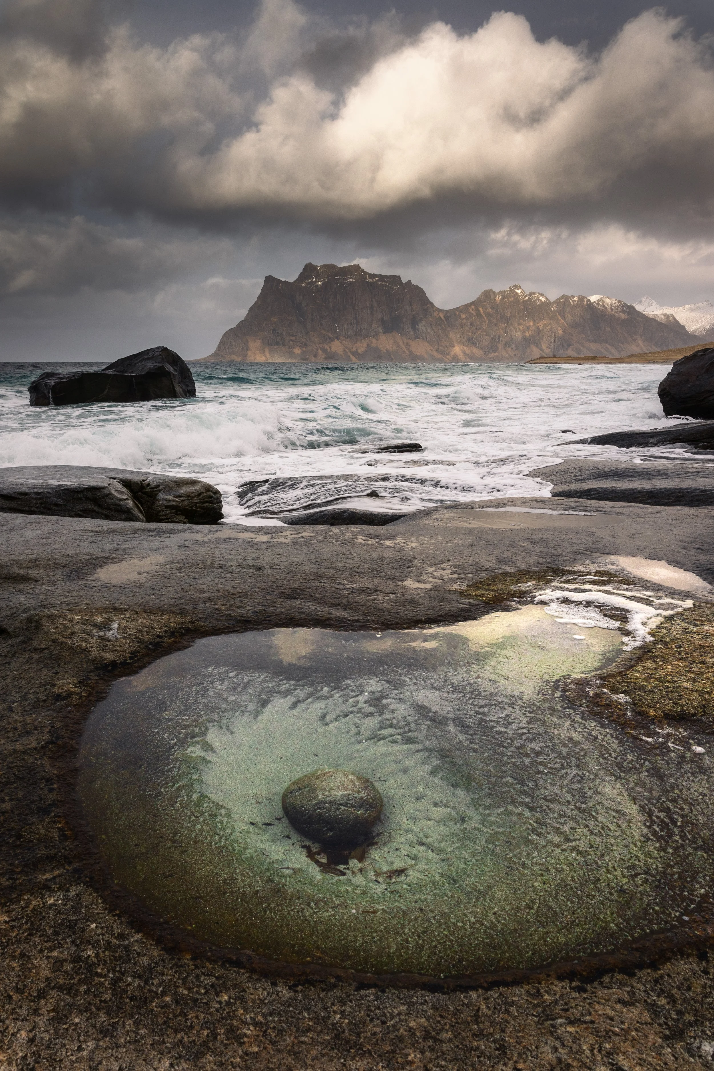 The Eye of Uttakleiv, a glacially scoured rock pool with a boulder at its centre, Lofoten, Norway — the mountain behind catching light through a break in the cloud.