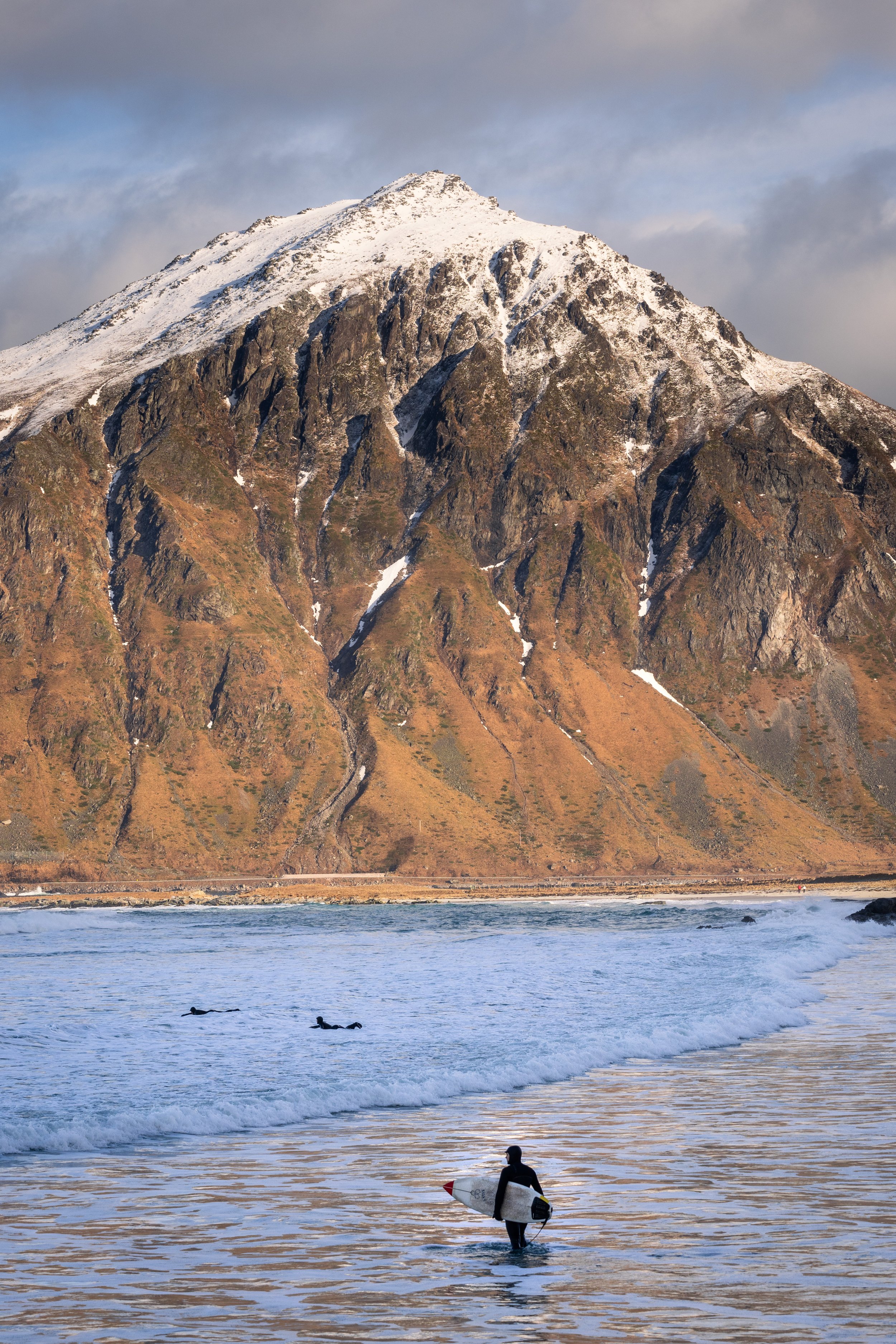 Arctic surfing. A surfer wading out beneath a snow-covered peak, in Lofoten, Norway — among the northernmost surf breaks in the world, ridden year-round above the Arctic Circle.