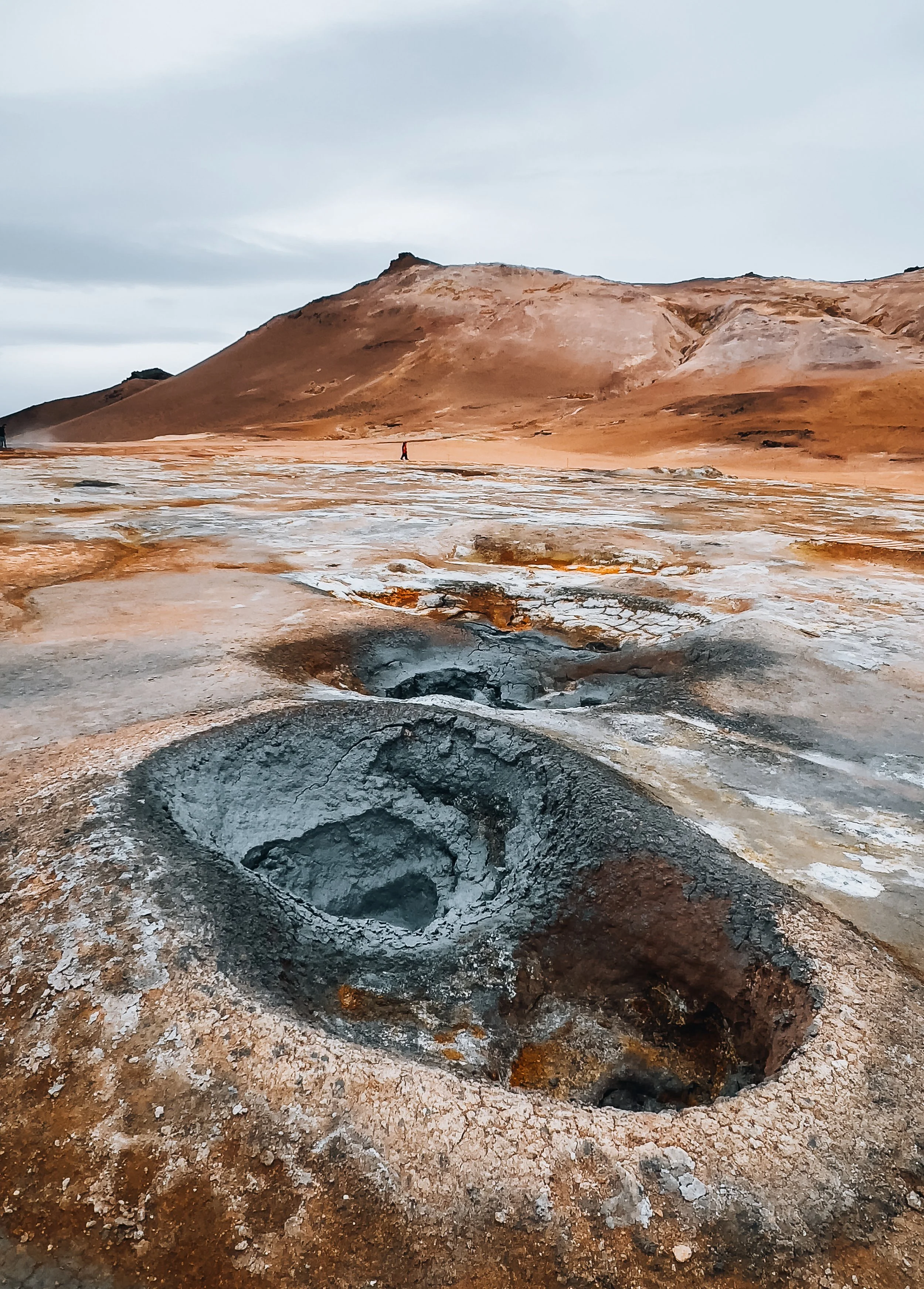 Bubbling grey mud pool rimmed with sulphur-stained crust below a rust-red ridge, Hverir geothermal field, north Iceland