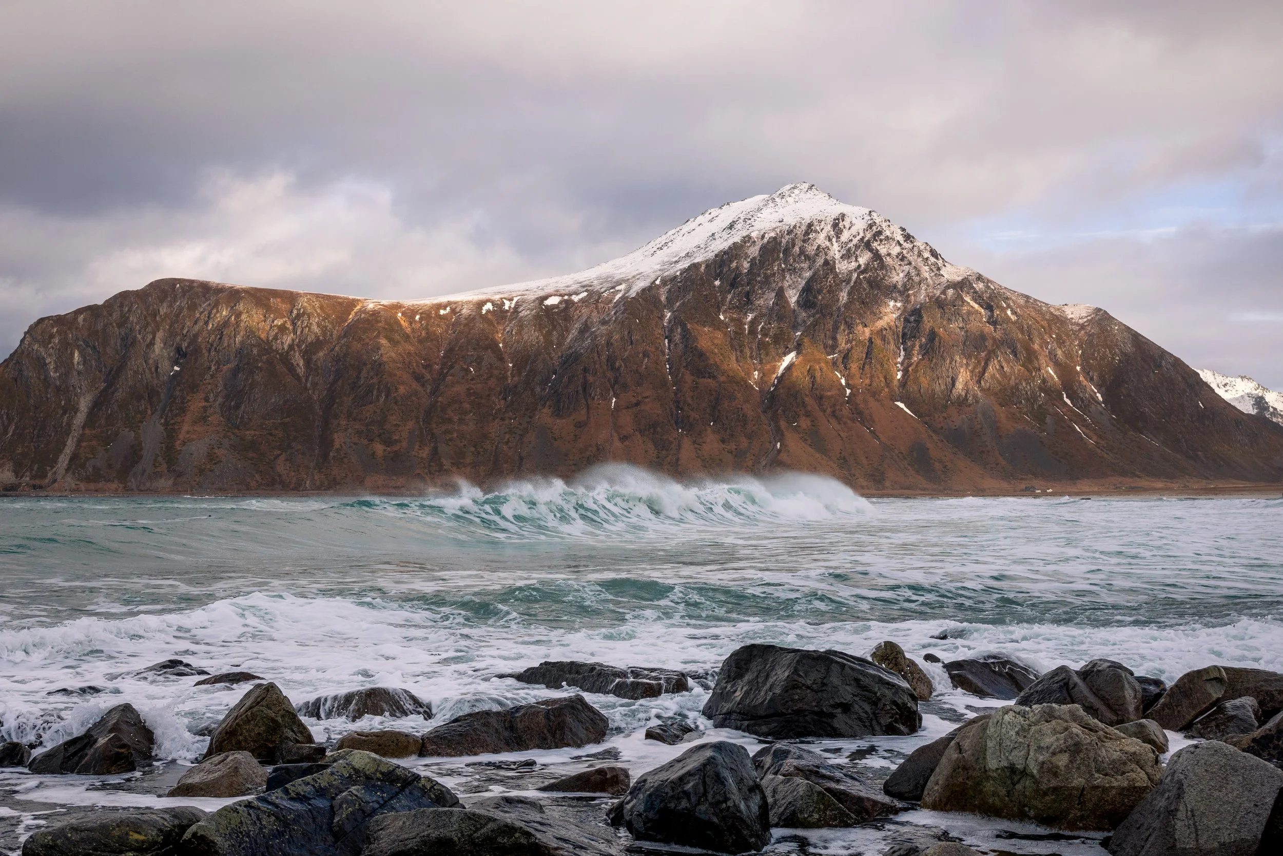 A wave breaking below a sunlit peak above a boulder shore, Lofoten, Norway — late light at this latitude runs for hours rather than minutes.