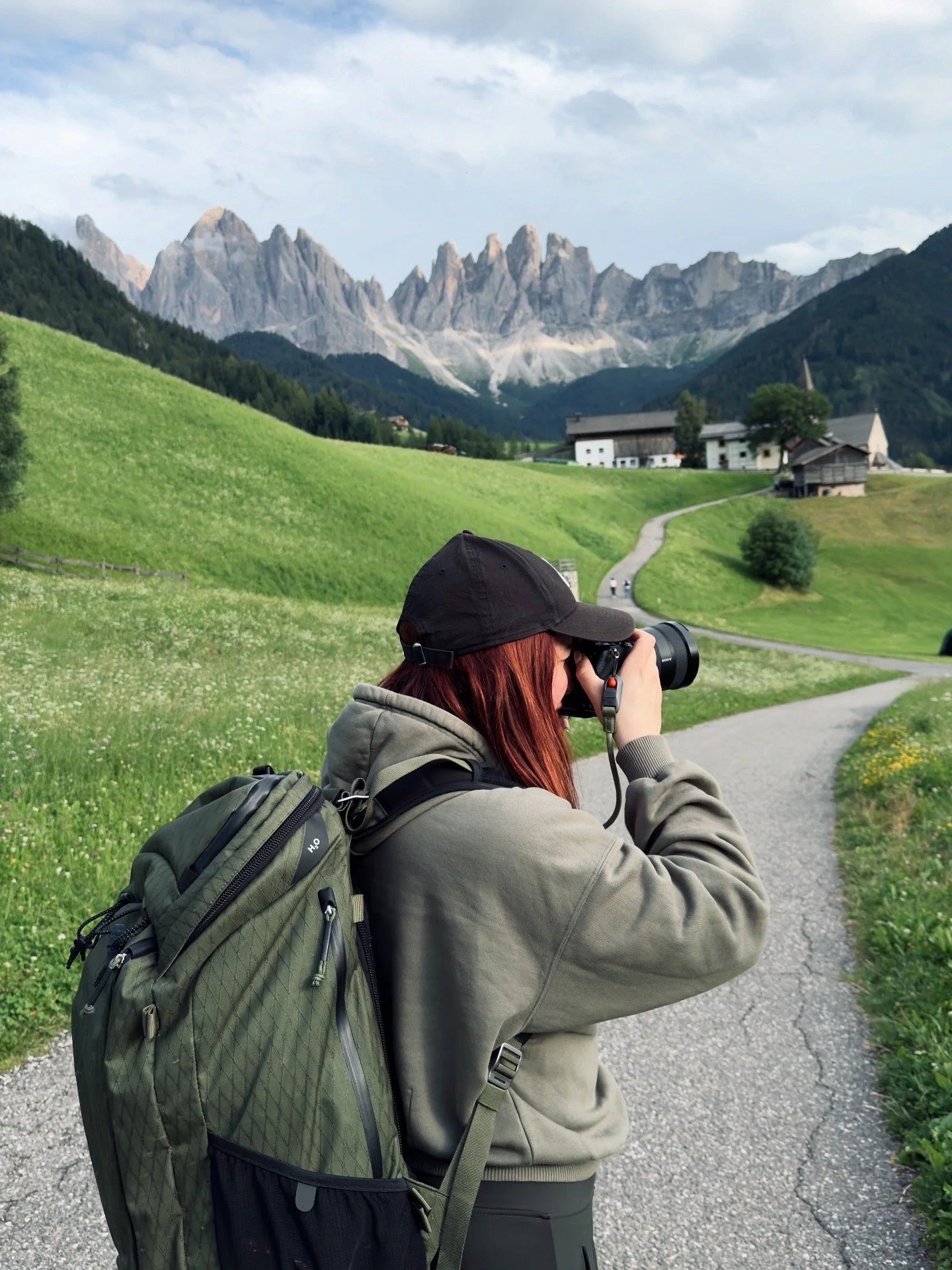 Vasiliki at Santa Maddalena in the Val di Funes, Italian Dolomites, camera raised to photograph the Odle group rising behind the village. The jagged limestone spires catch light on their upper faces with cloud gathering around them, while below the v