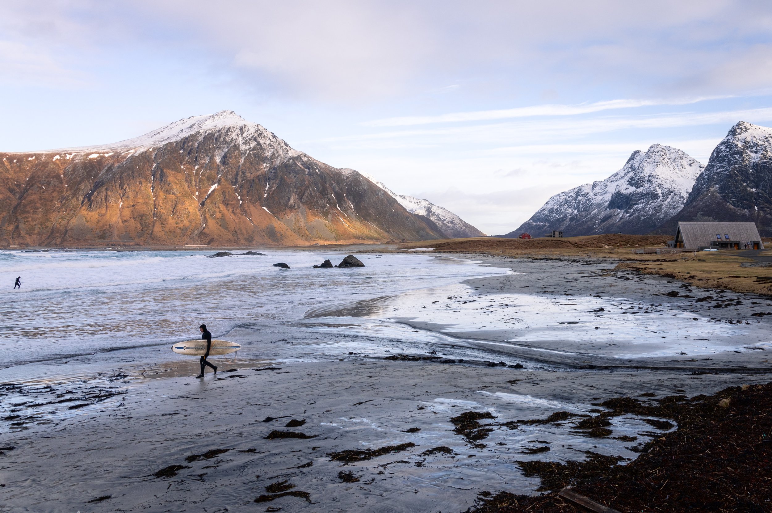 A surfer crossing the tidal flats with a board under one arm, Lofoten, Norway — low winter sun on the ridge, the whole beach empty behind them.