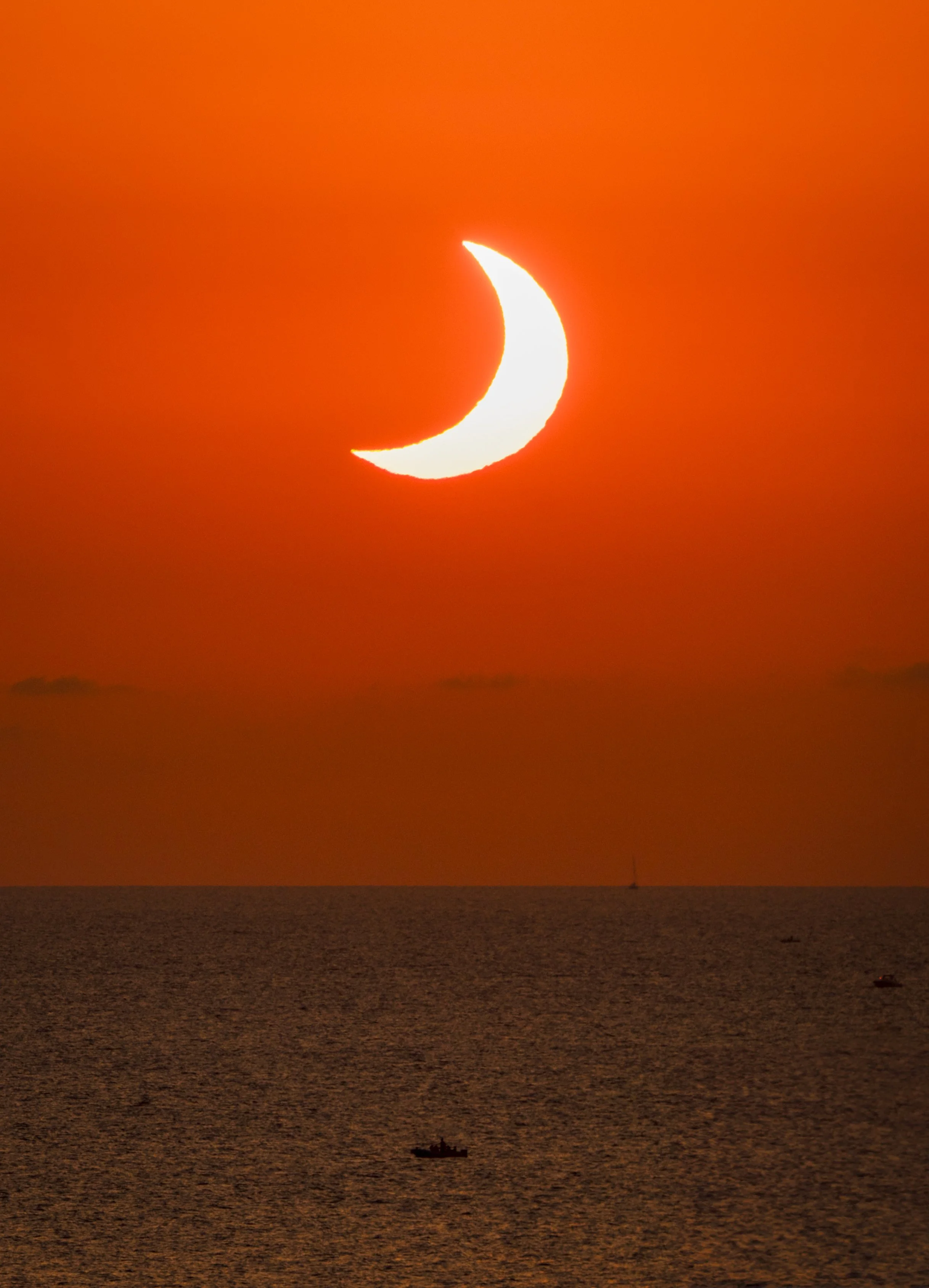 A late stage of the partial phase on 12 August 2026 over Ibiza, the sun worn down to a thin crescent tilted on its side. Shot long, so the sun sits unusually large in the frame against a sky burned to deep red-orange, with the Mediterranean below it 