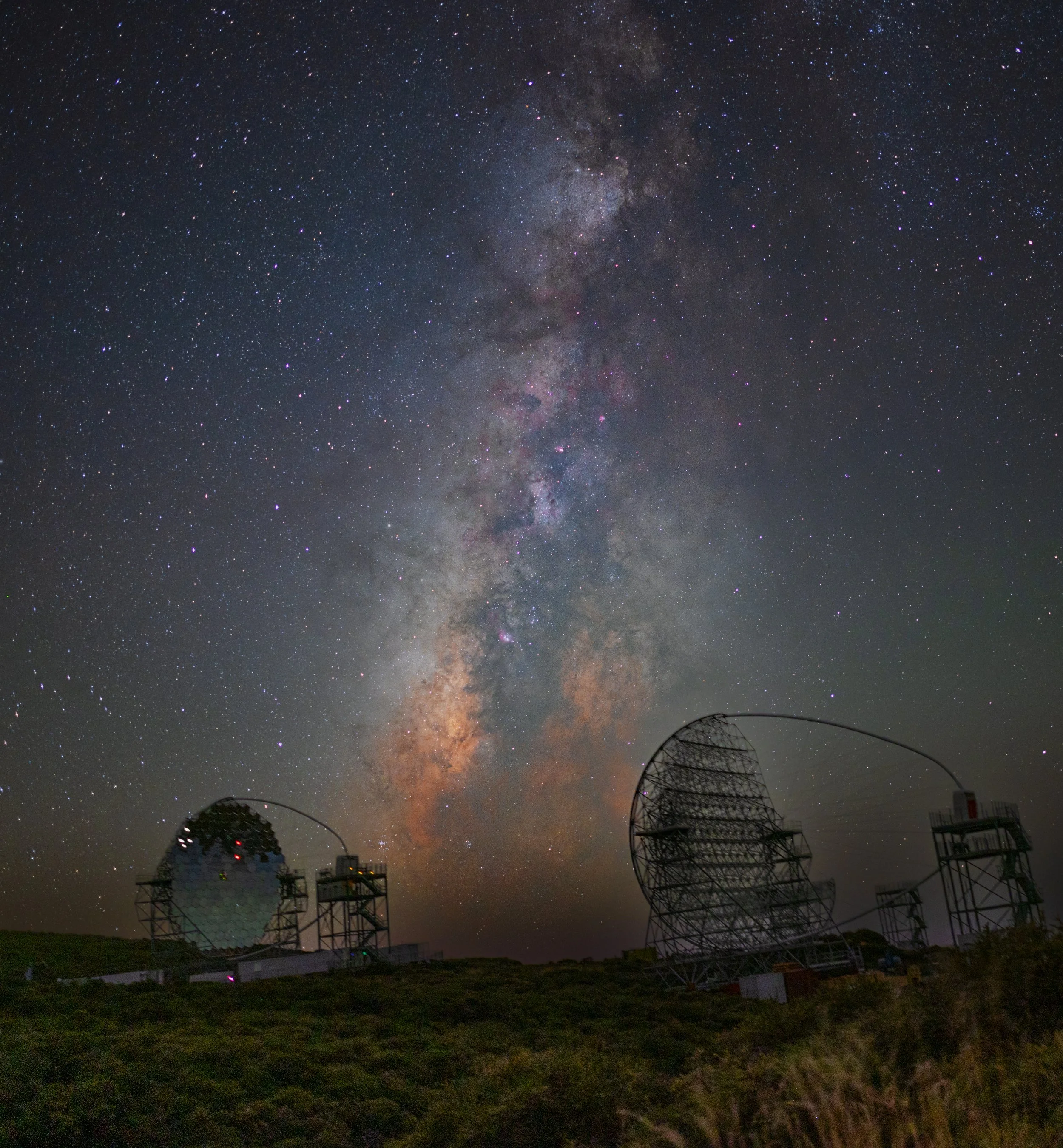 The Milky Way core above the MAGIC gamma-ray telescopes at Roque de los Muchachos Observatory, La Palma — their segmented mirrors catch the faint blue flashes produced when gamma rays from distant galaxies strike the upper atmosphere, which is why th
